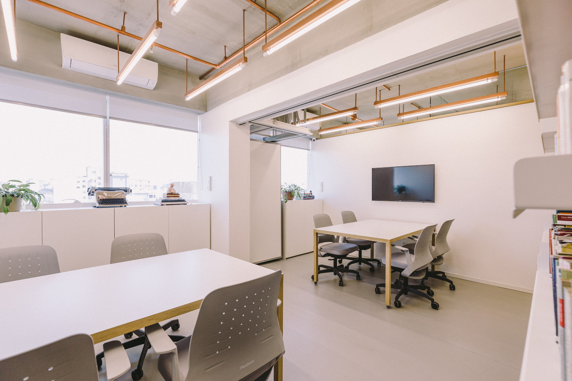 Meeting room with two white tables, grey perforated chairs and copper-framed tube lights, daylight pouring through the window