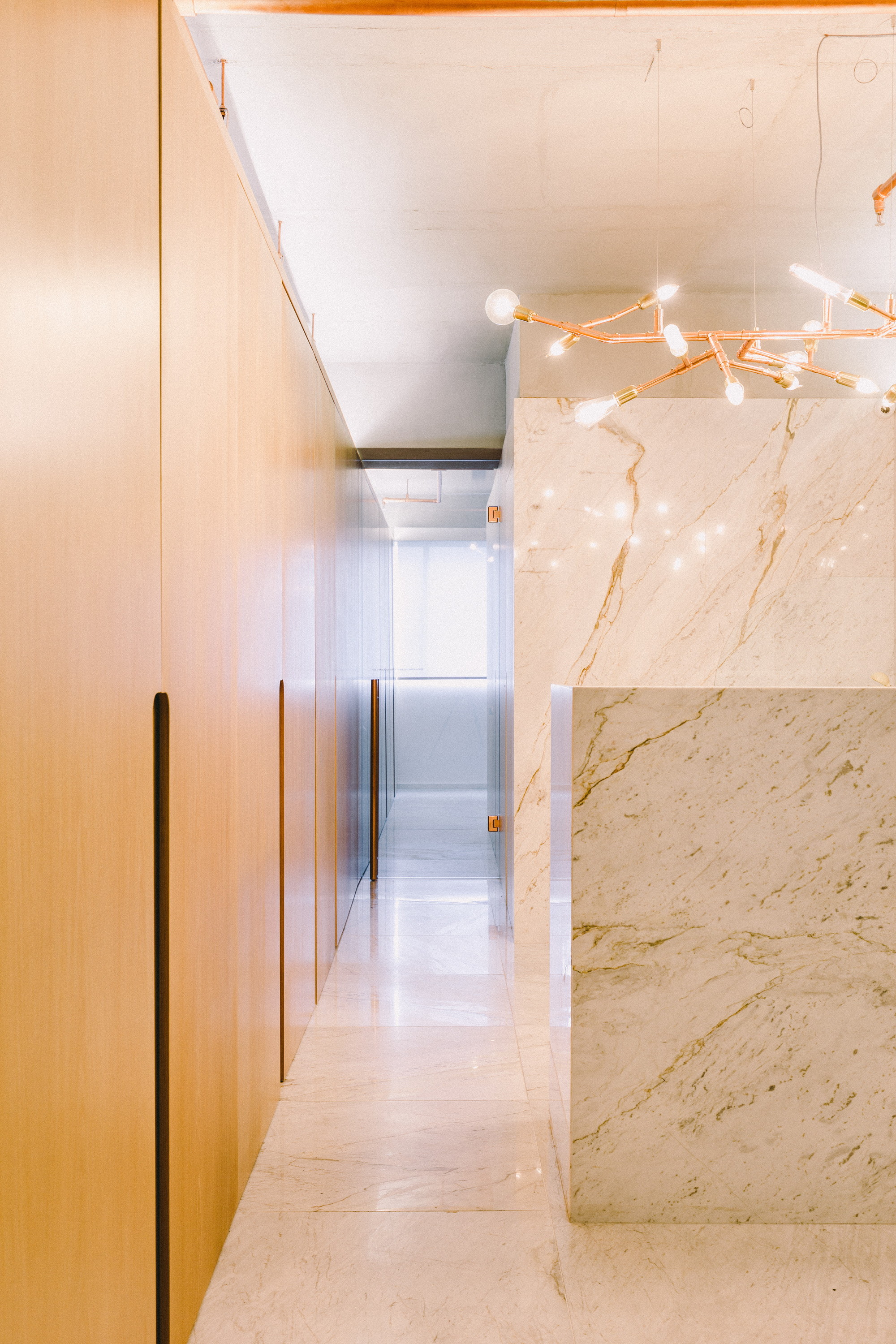 Corridor lined with oak cabinet doors and recessed pulls, ending beside a marble-clad block and a branching copper chandelier