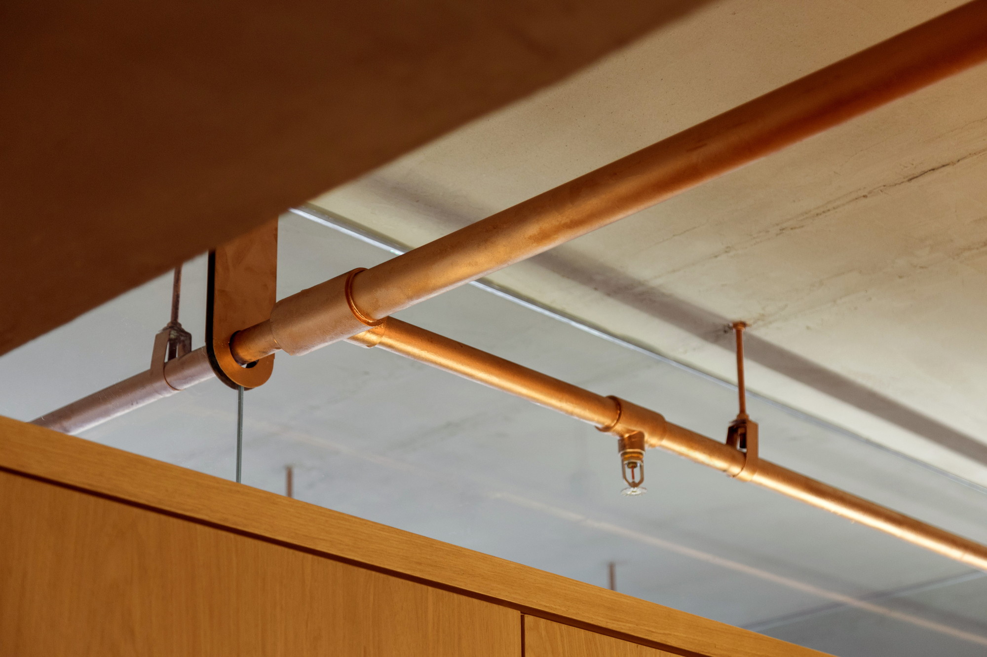 Close-up of a copper sprinkler pipe with a bracket and sprinkler head above an oak panel edge