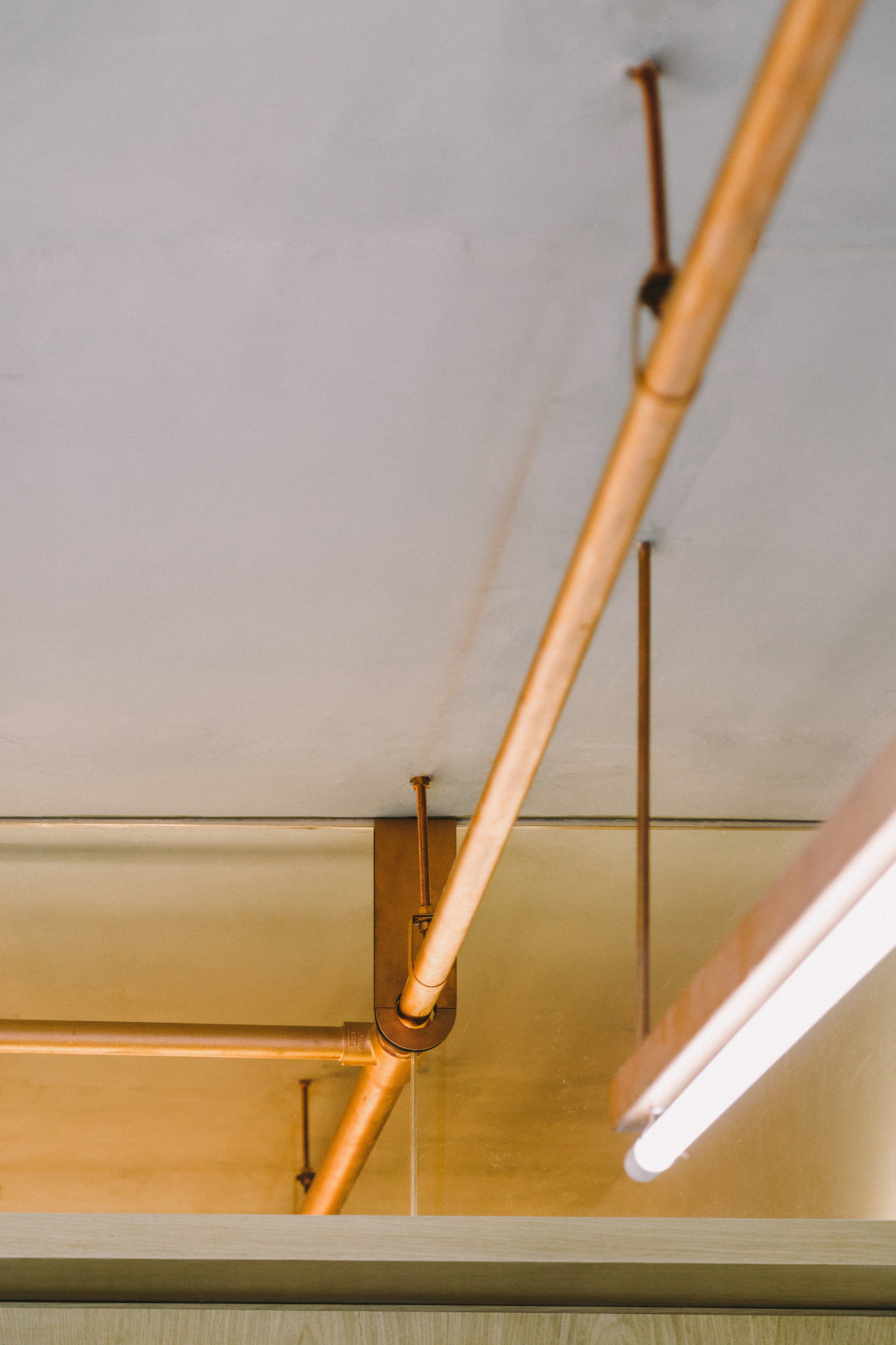 Looking up at a diagonal copper pipe joined to a bracket under a concrete ceiling, with a tube light at right