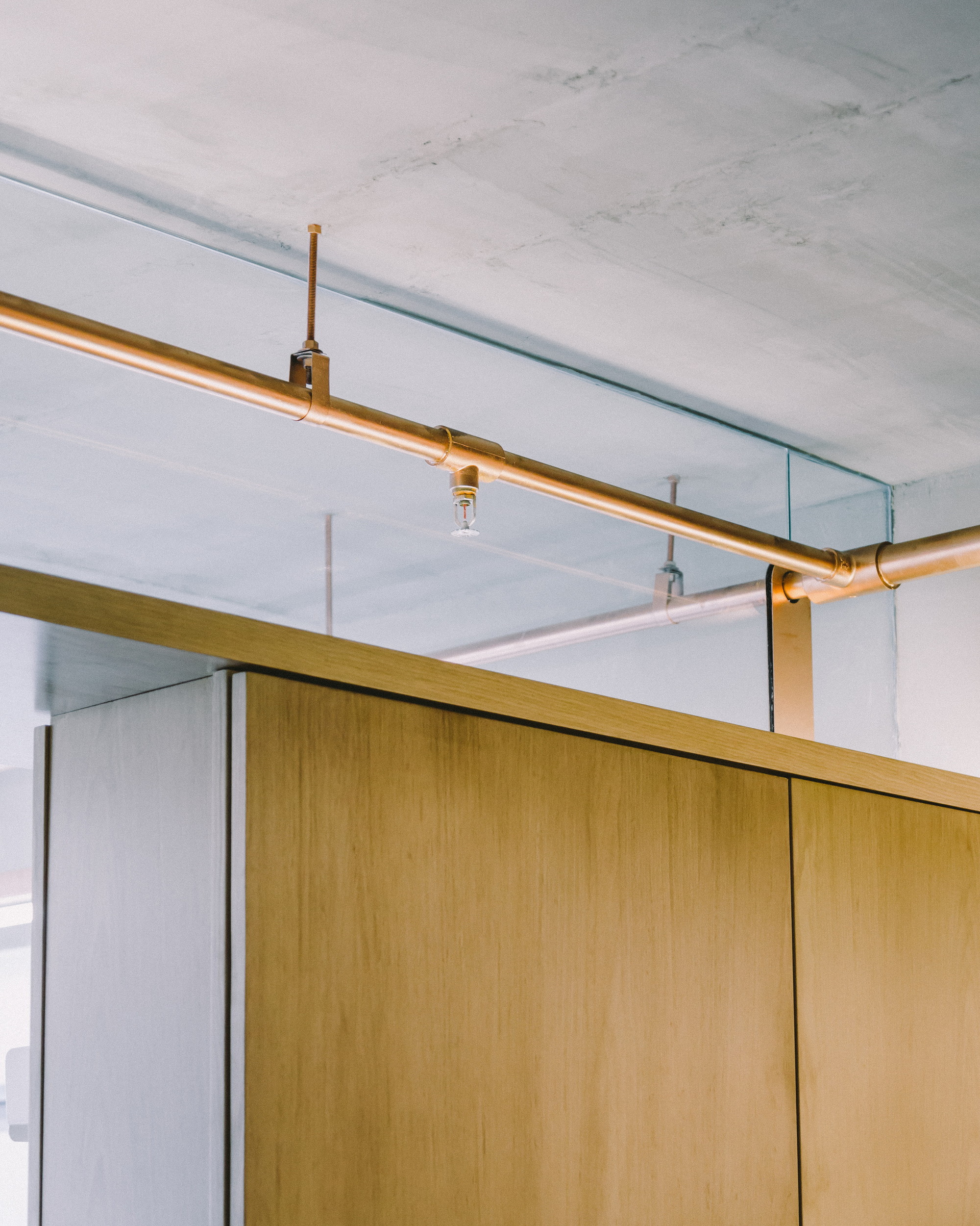 Copper sprinkler line running along a pale concrete ceiling above oak cabinets, with a glass gap between ceiling and joinery