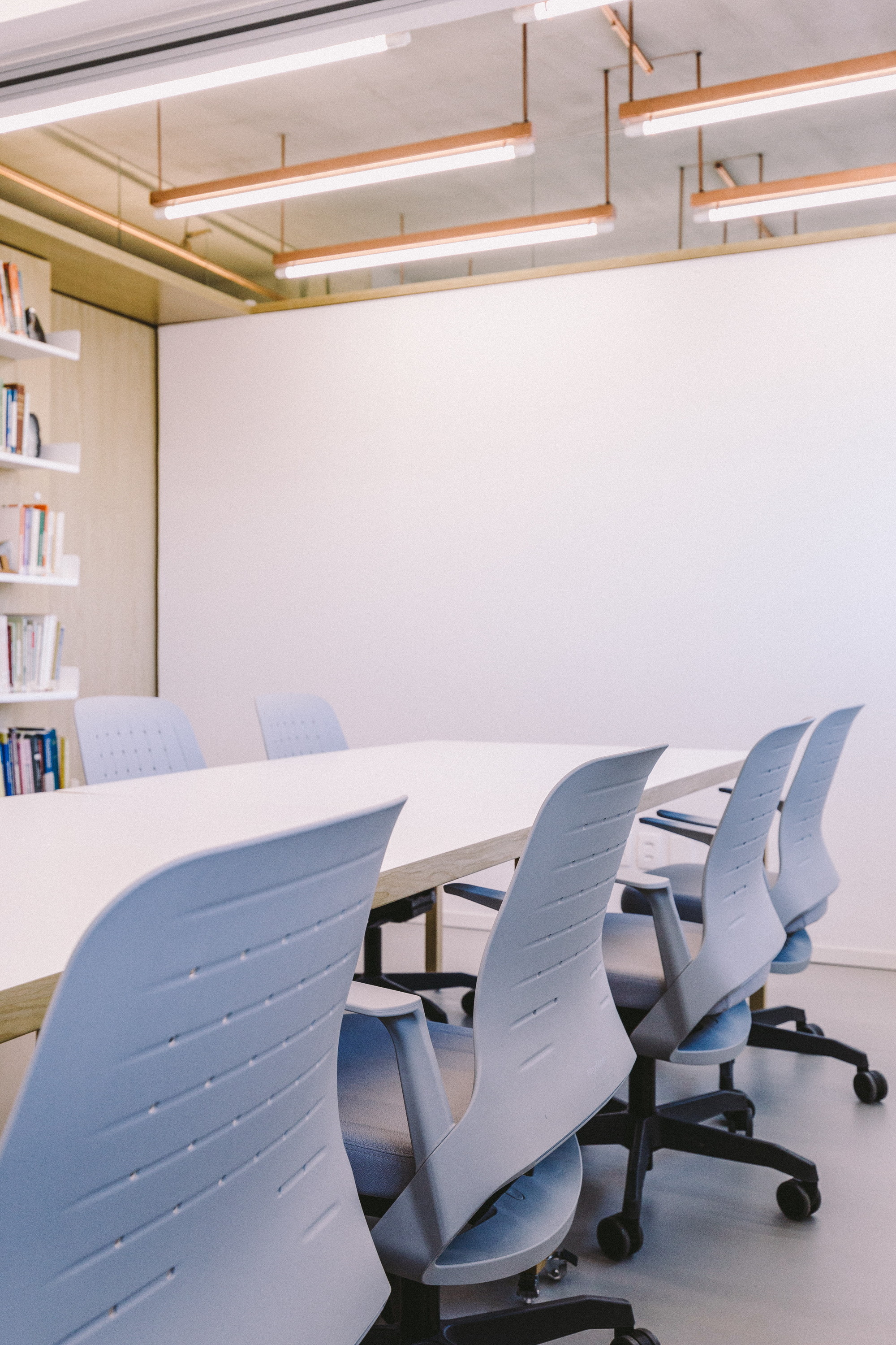 Meeting table with pale grey task chairs beside a wall of oak shelving, under copper-framed linear light fixtures