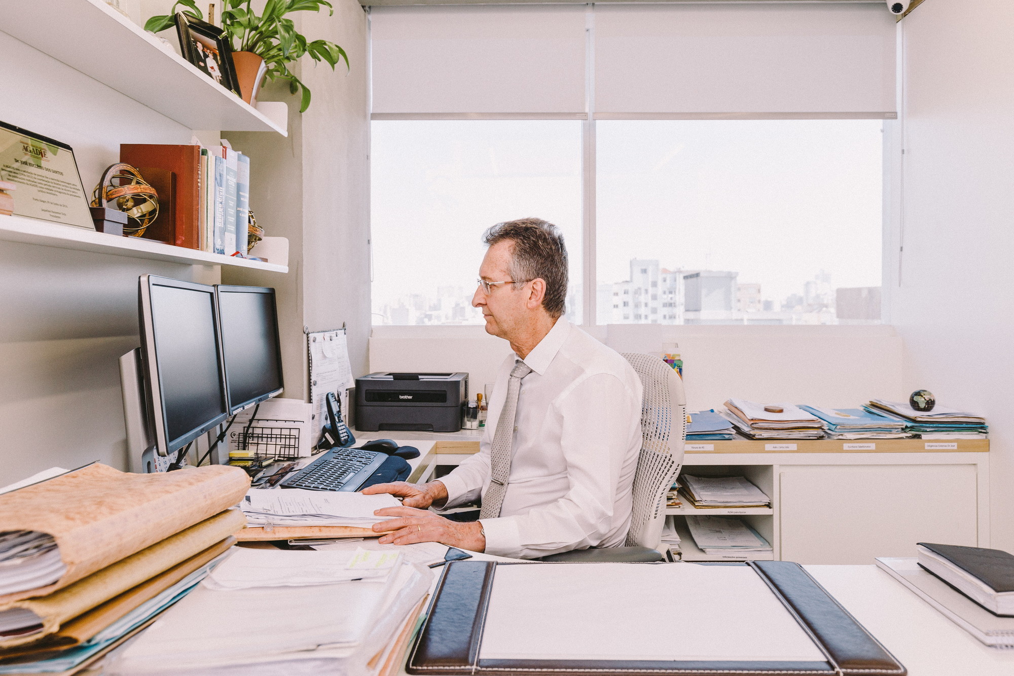 Man in a white shirt working at a desk with two monitors beside a window overlooking distant buildings