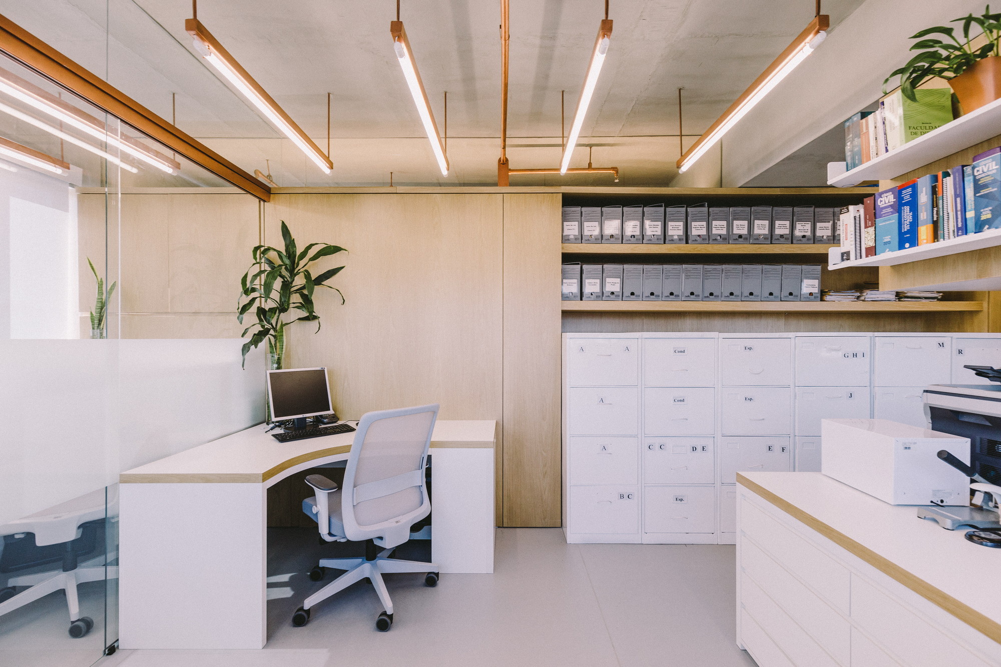 Workstation with curved white desk and mesh chair beside a glass partition, with filing cabinets under copper light fittings