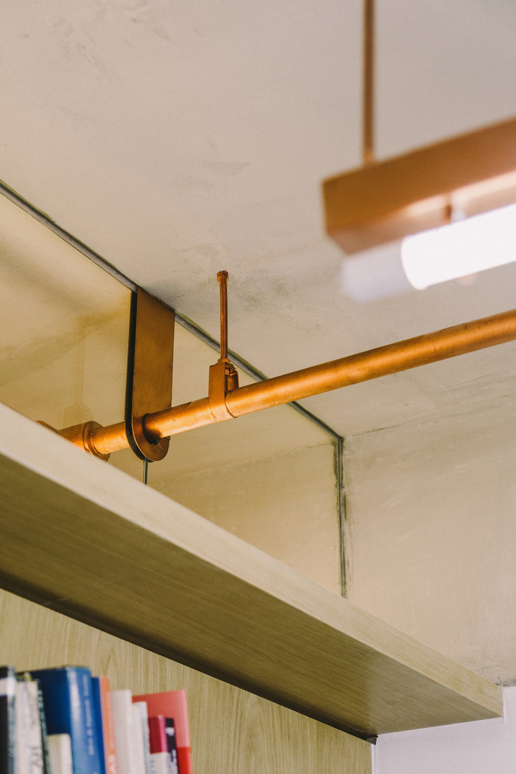 Close-up of a copper pipe and bracket running above an oak shelf, with a light fixture beside the concrete ceiling