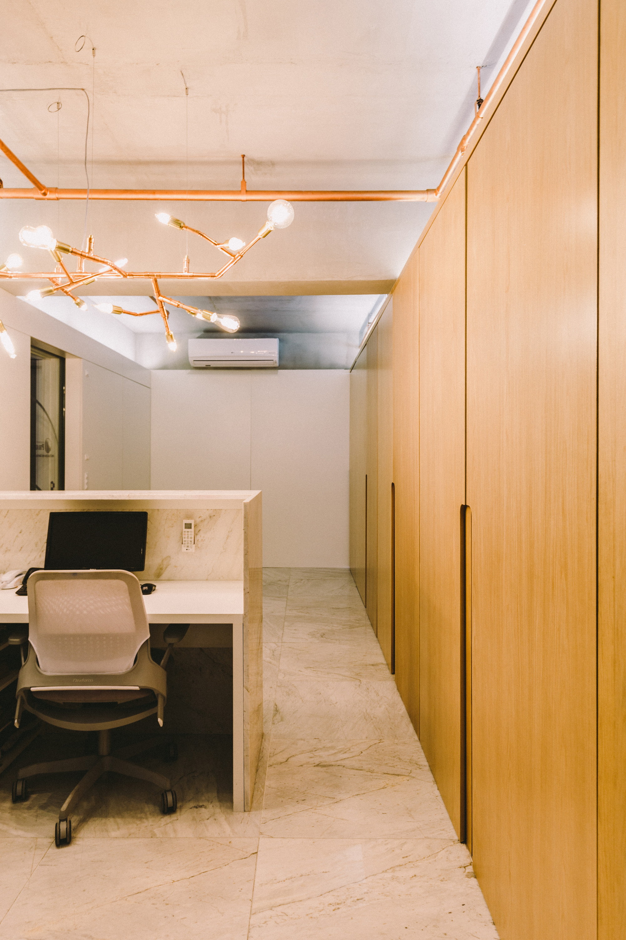 Reception desk with a mesh chair facing a copper branch chandelier, alongside a long oak panel wall in soft daylight