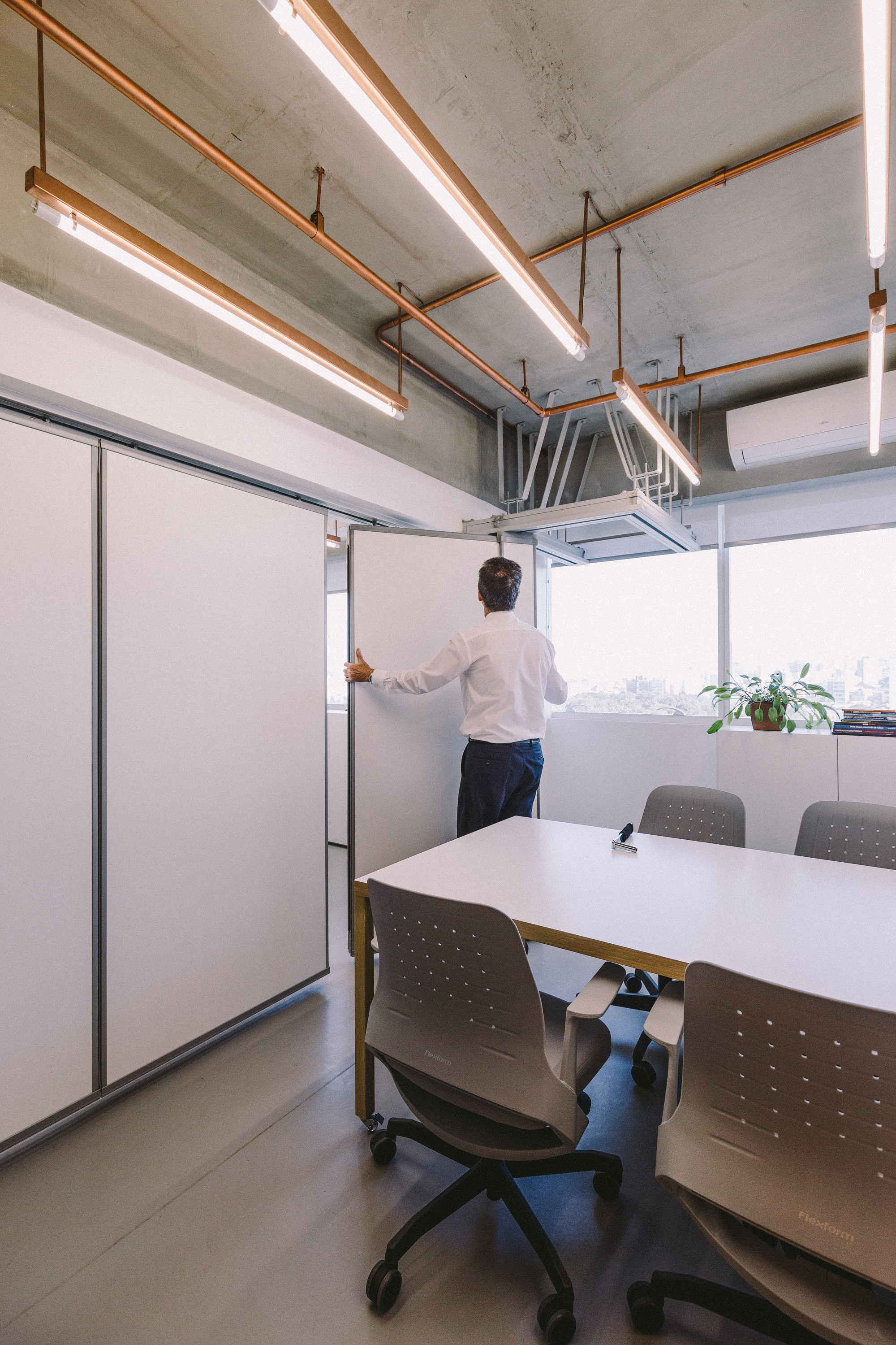 Man opening a tall white folding partition beside a meeting table, with window light falling on a potted plant
