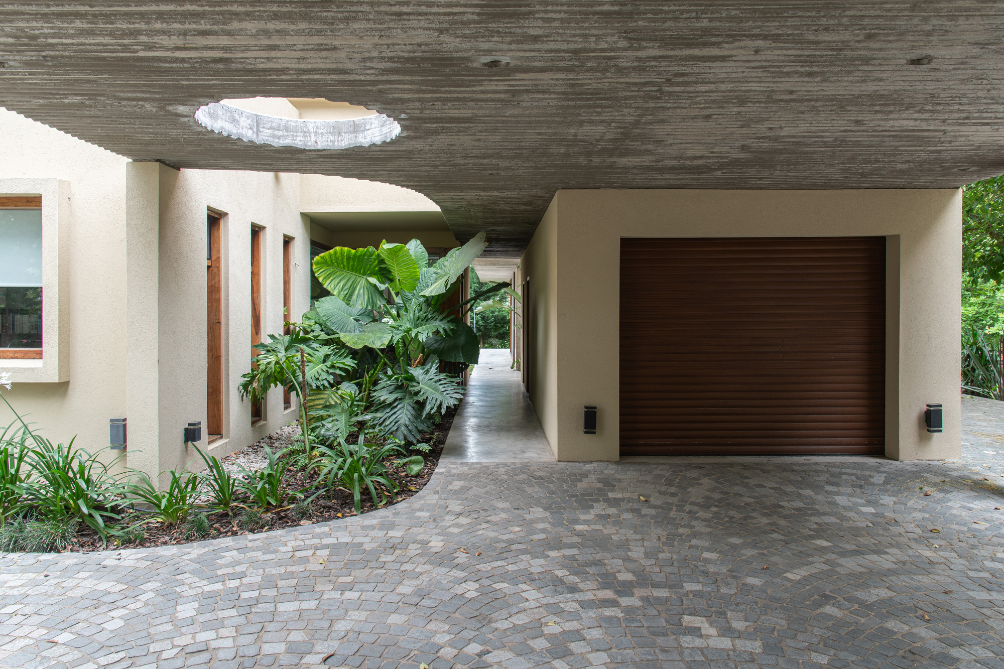 Board-formed concrete carport soffit with a round skylight above a curved planting bed of large-leaf tropical plants and a brown roller garage door