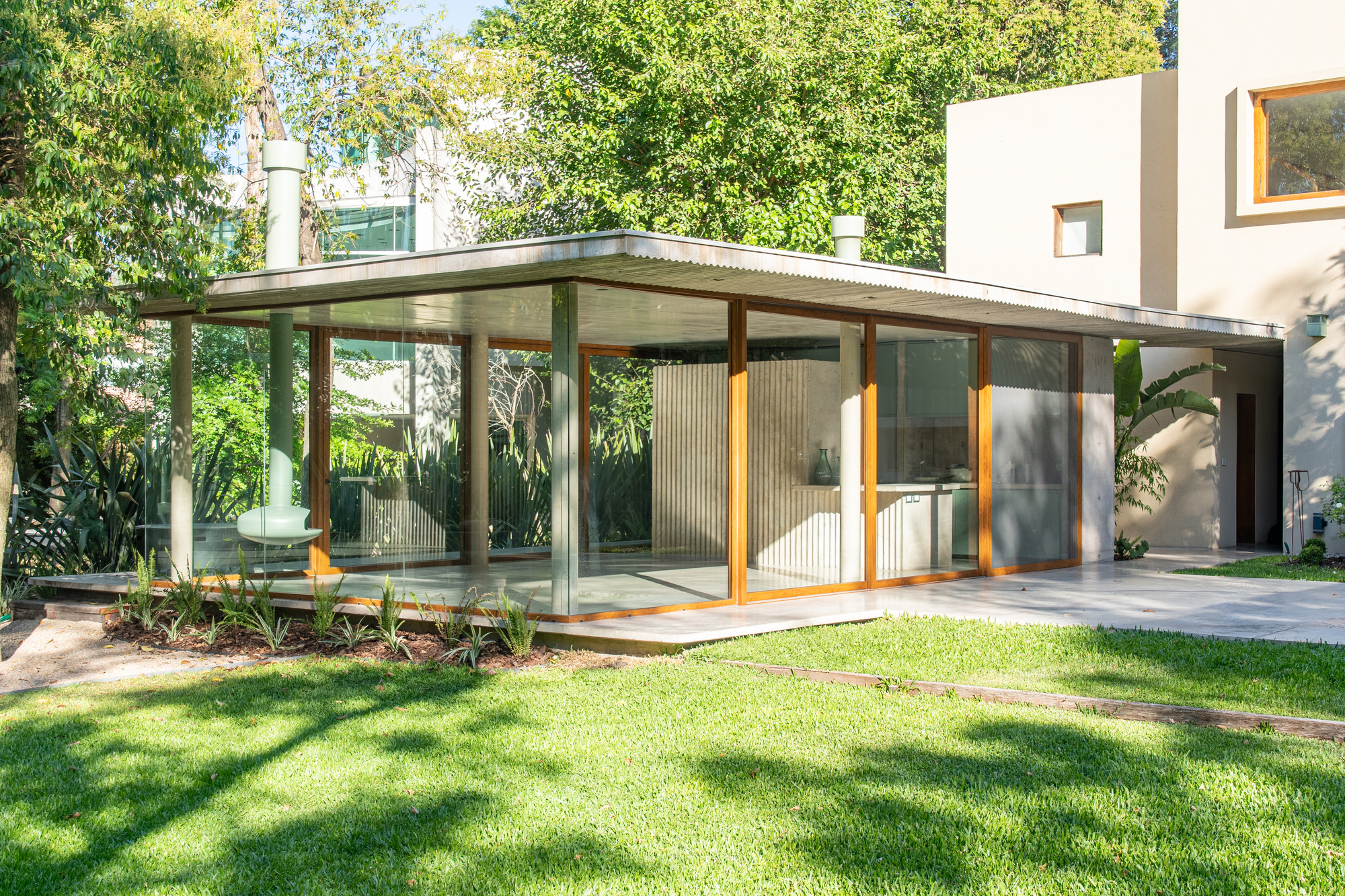 Glass-walled pavilion with timber frames and a thin flat roof, seen across a sunlit lawn dappled by tree shadows