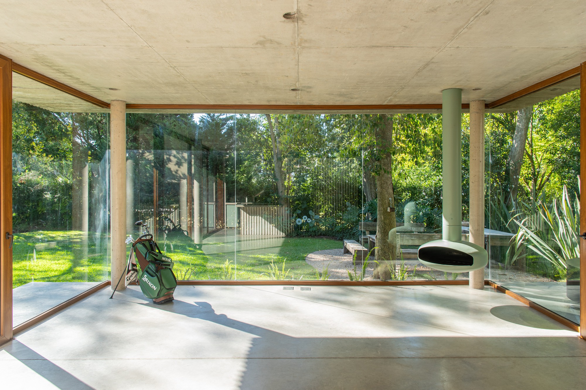 Inside the glazed pavilion, a suspended green fireplace hangs beside slim concrete columns, with a golf bag on the floor and garden trees beyond