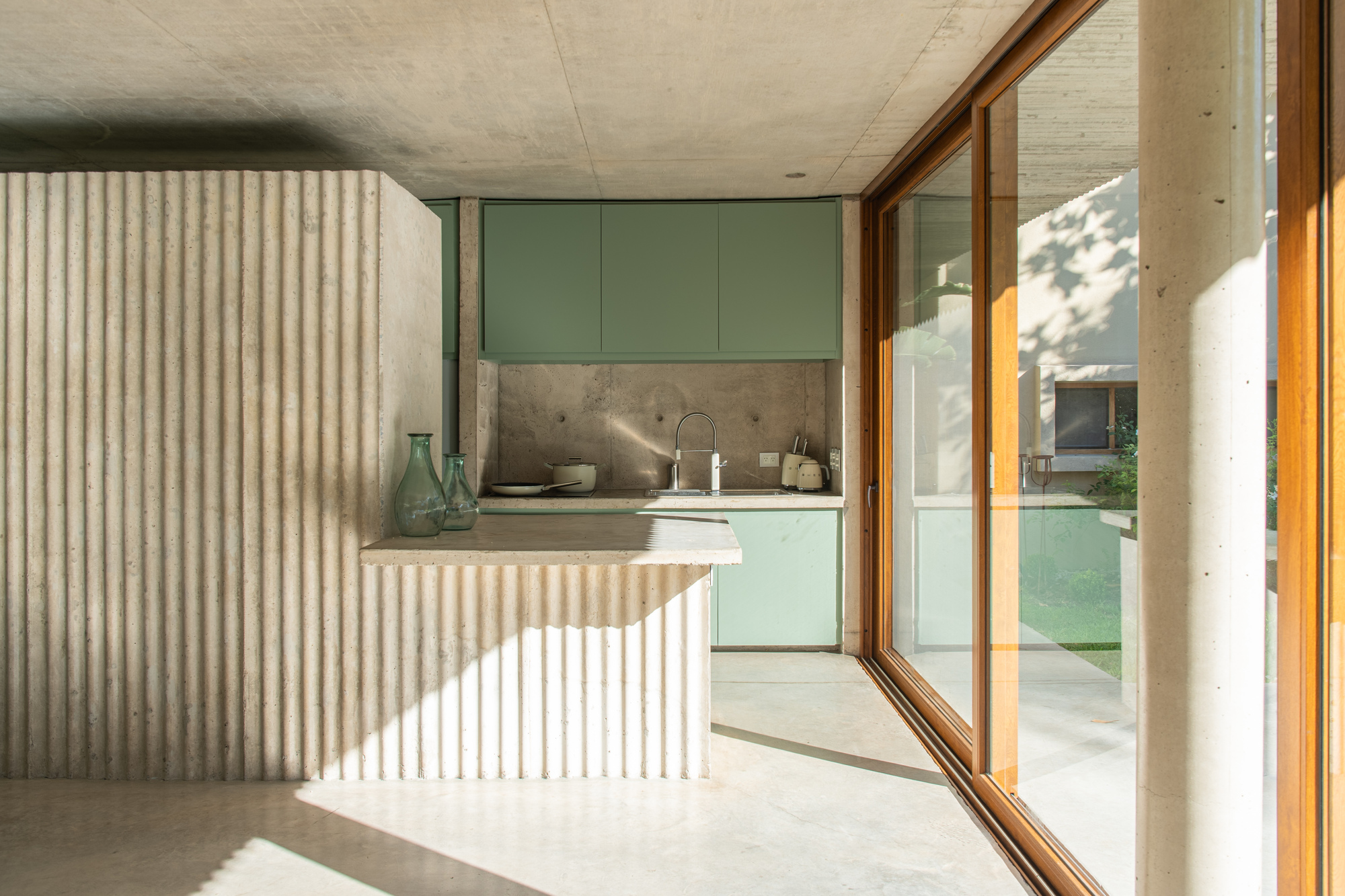 Kitchen corner with a ribbed concrete counter, sage green cabinets and glass vases, lit by low sun through timber-framed glazing