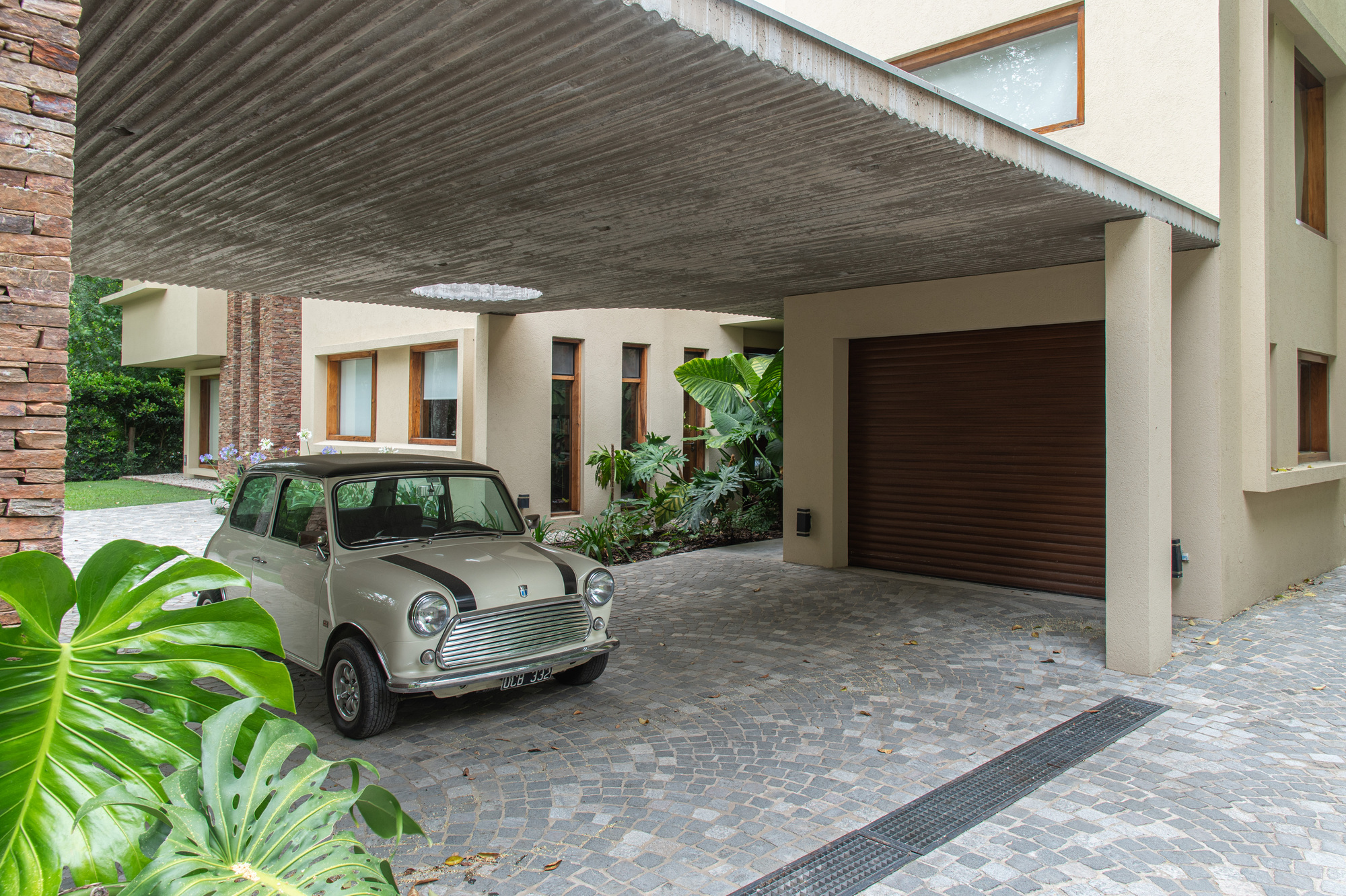 Cream vintage mini parked on cobblestone paving under a board-formed concrete canopy, with a monstera leaf in the foreground and a brick pier at left