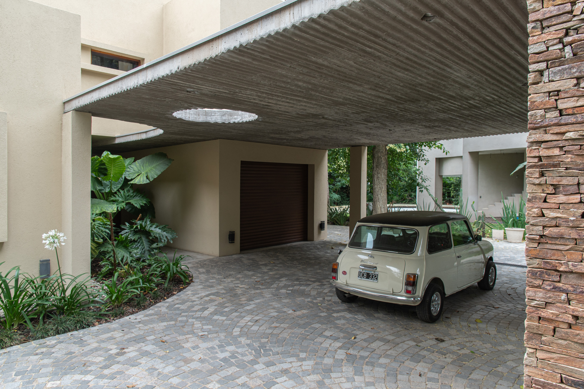 Rear view of a cream vintage car beneath the concrete carport canopy, with a round skylight, white agapanthus and a stacked stone pier at right