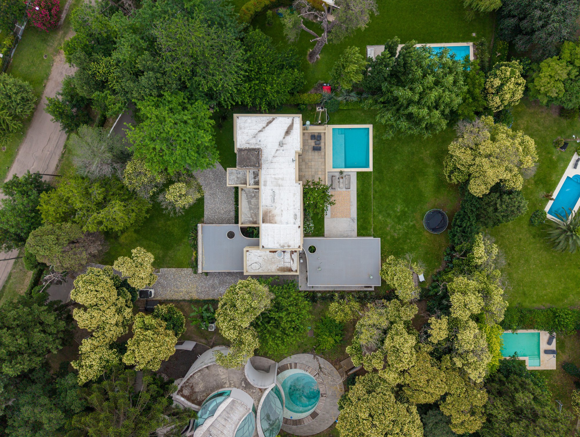 Aerial view of flat-roofed house volumes among dense tree canopy, with a turquoise pool and neighbouring gardens