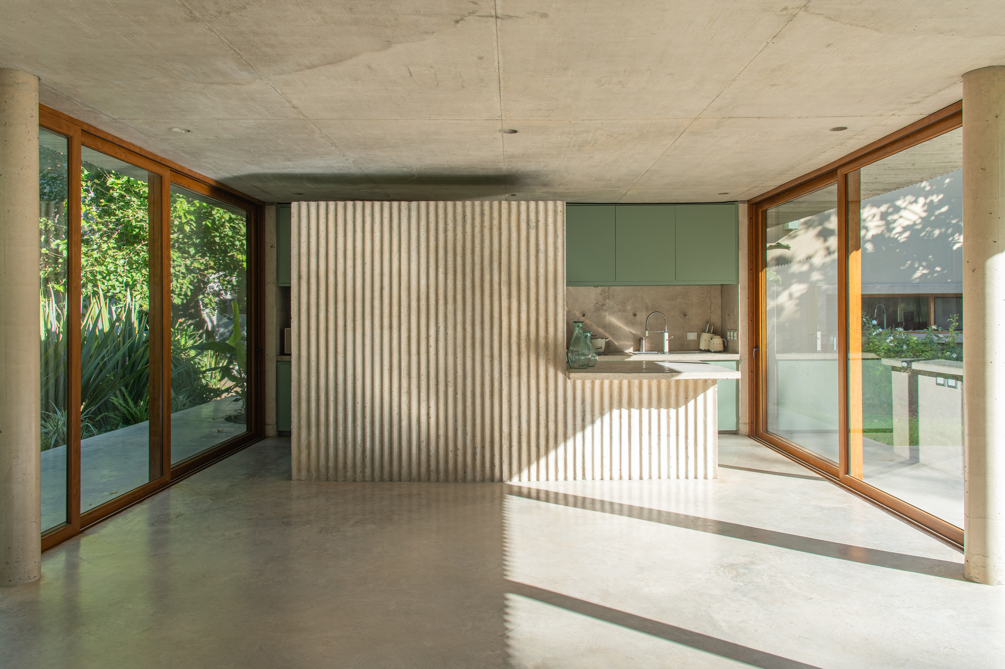 Open interior with a fluted travertine-clad block, green kitchen cabinets and timber-framed glass doors, with low sun casting long shadows on the polished floor