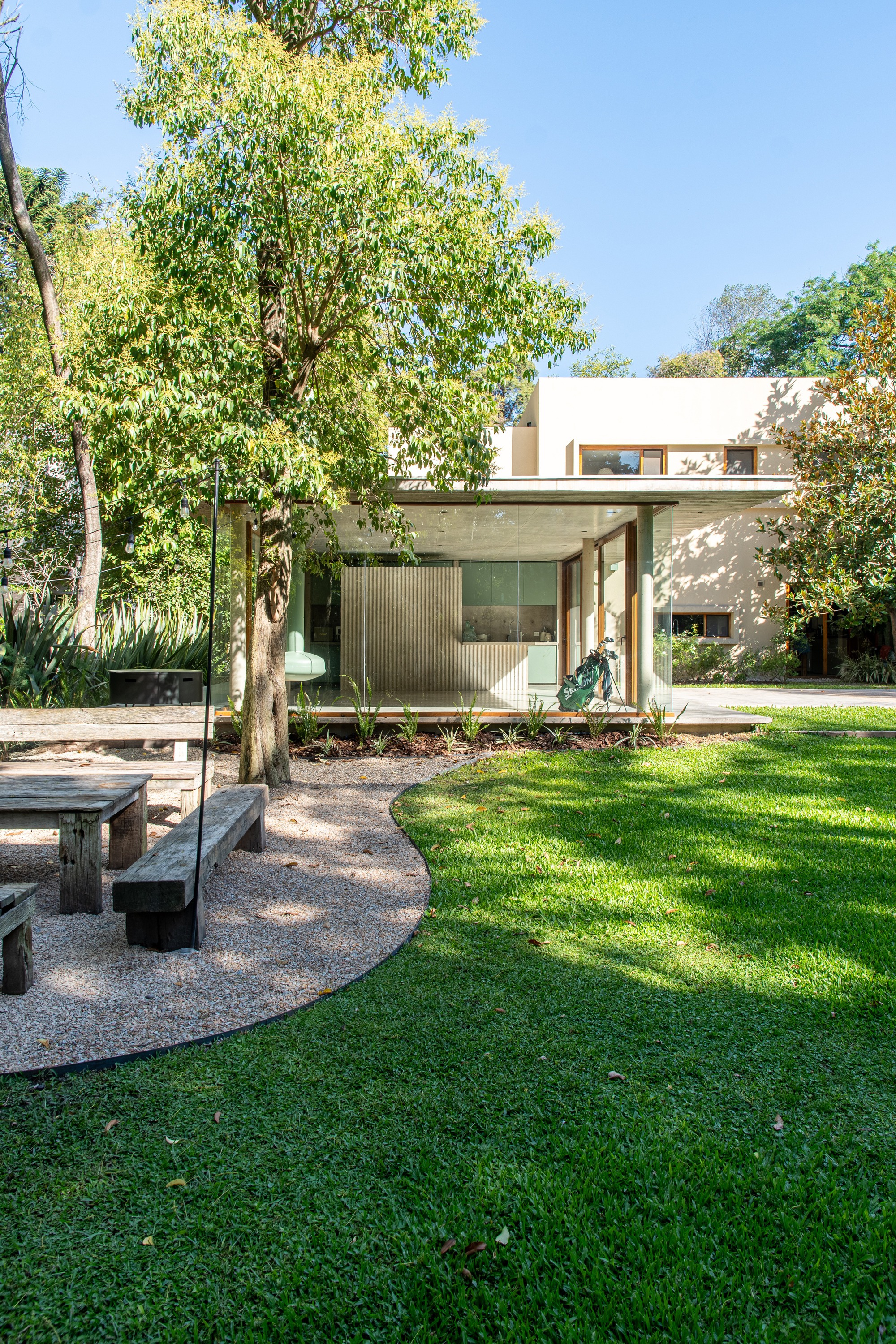 Glazed pavilion beside the two-storey house, seen across a lawn and gravel path with timber benches under a leafy tree