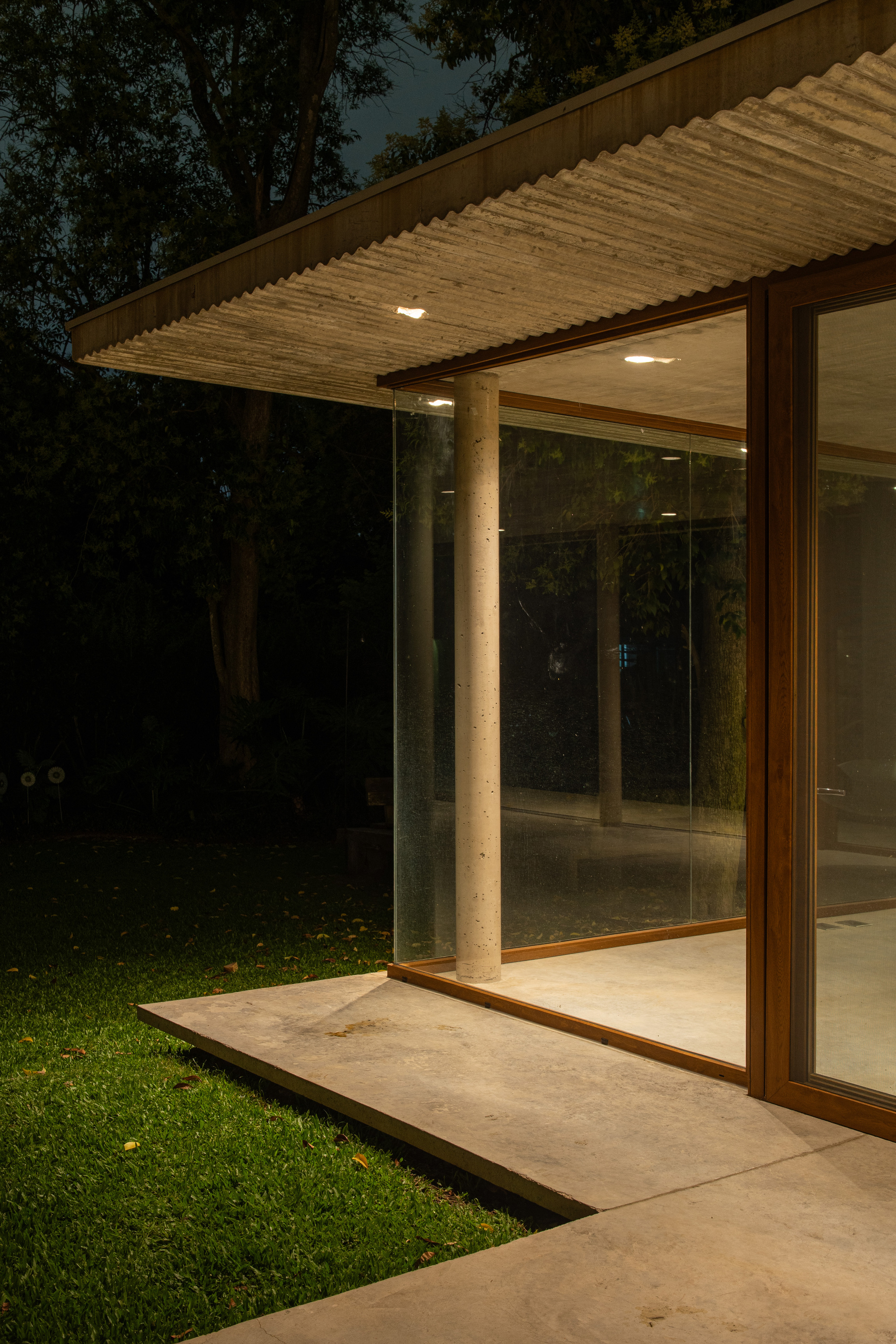 Night view of a glazed corner with a round concrete column under a corrugated roof, lawn lit in the foreground