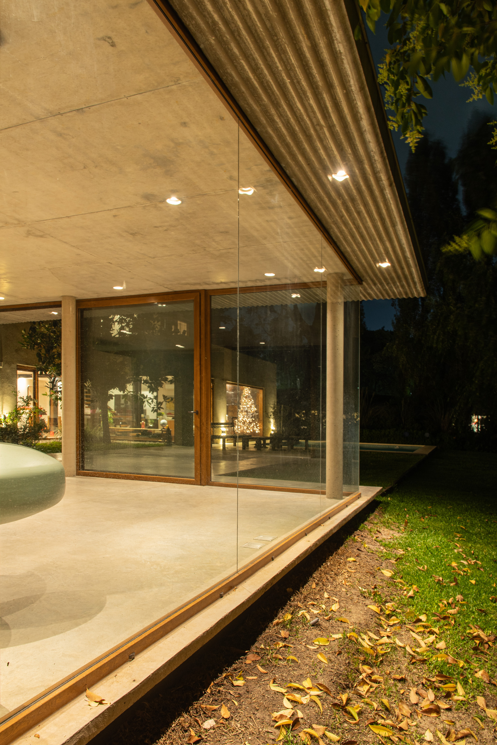 Glass corner and concrete column under a corrugated roof at dusk, fallen leaves on the ground and a lit tree inside