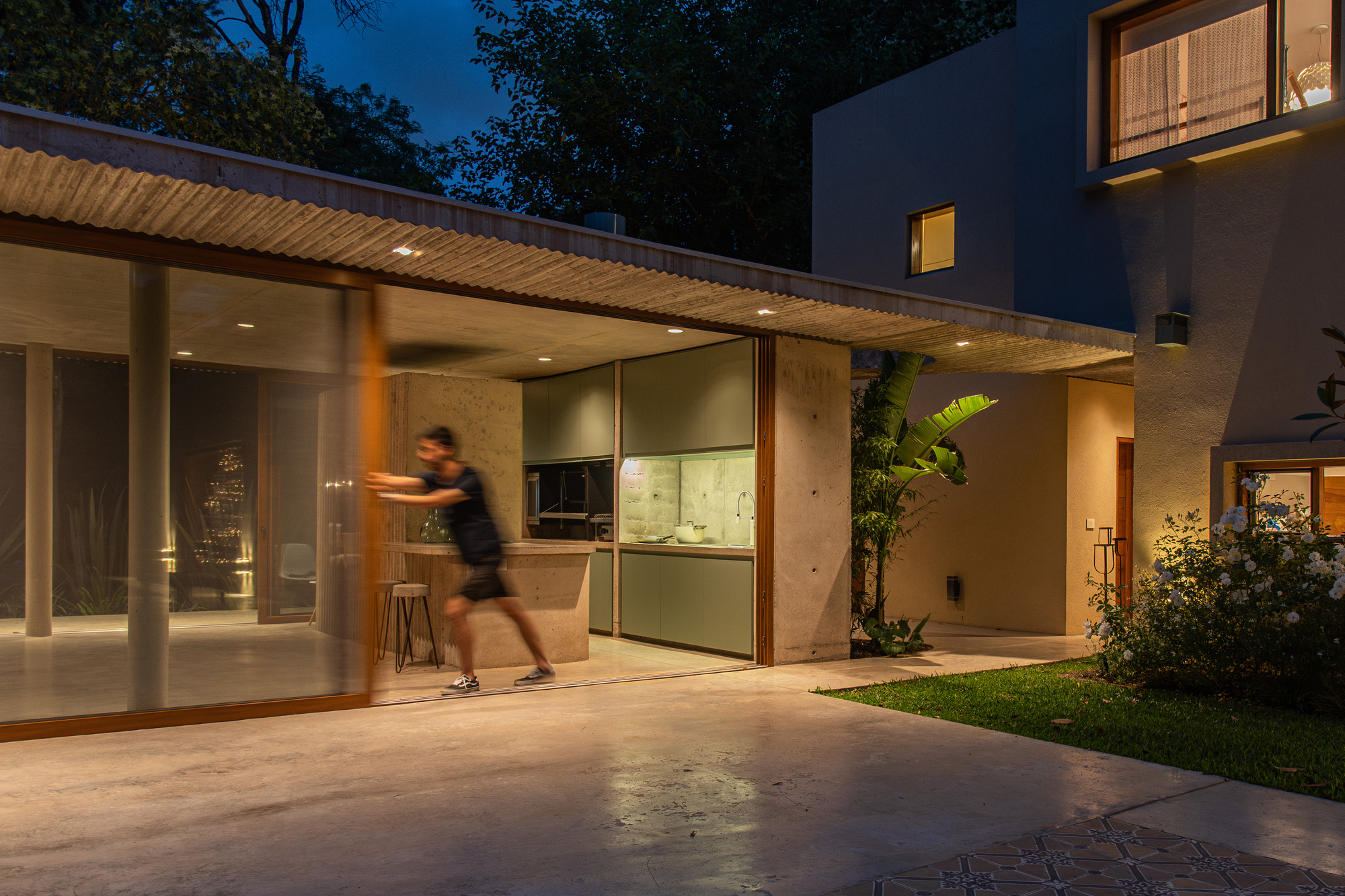Person pushing open a sliding glass door at dusk beside an open kitchen under a corrugated concrete roof