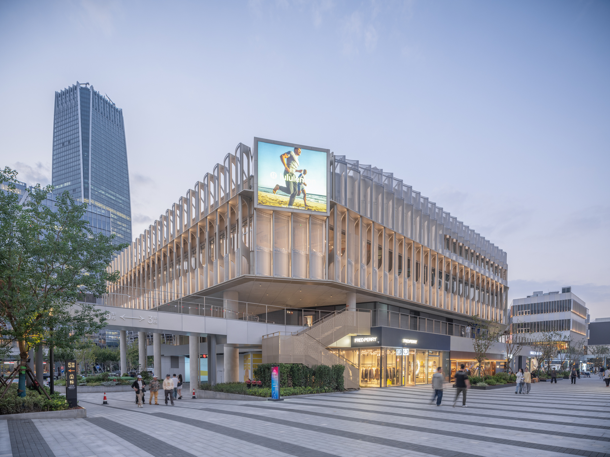 Corner of a multi-storey retail building wrapped in white vertical fins, with an exterior stair and pedestrians crossing the paved plaza at dusk