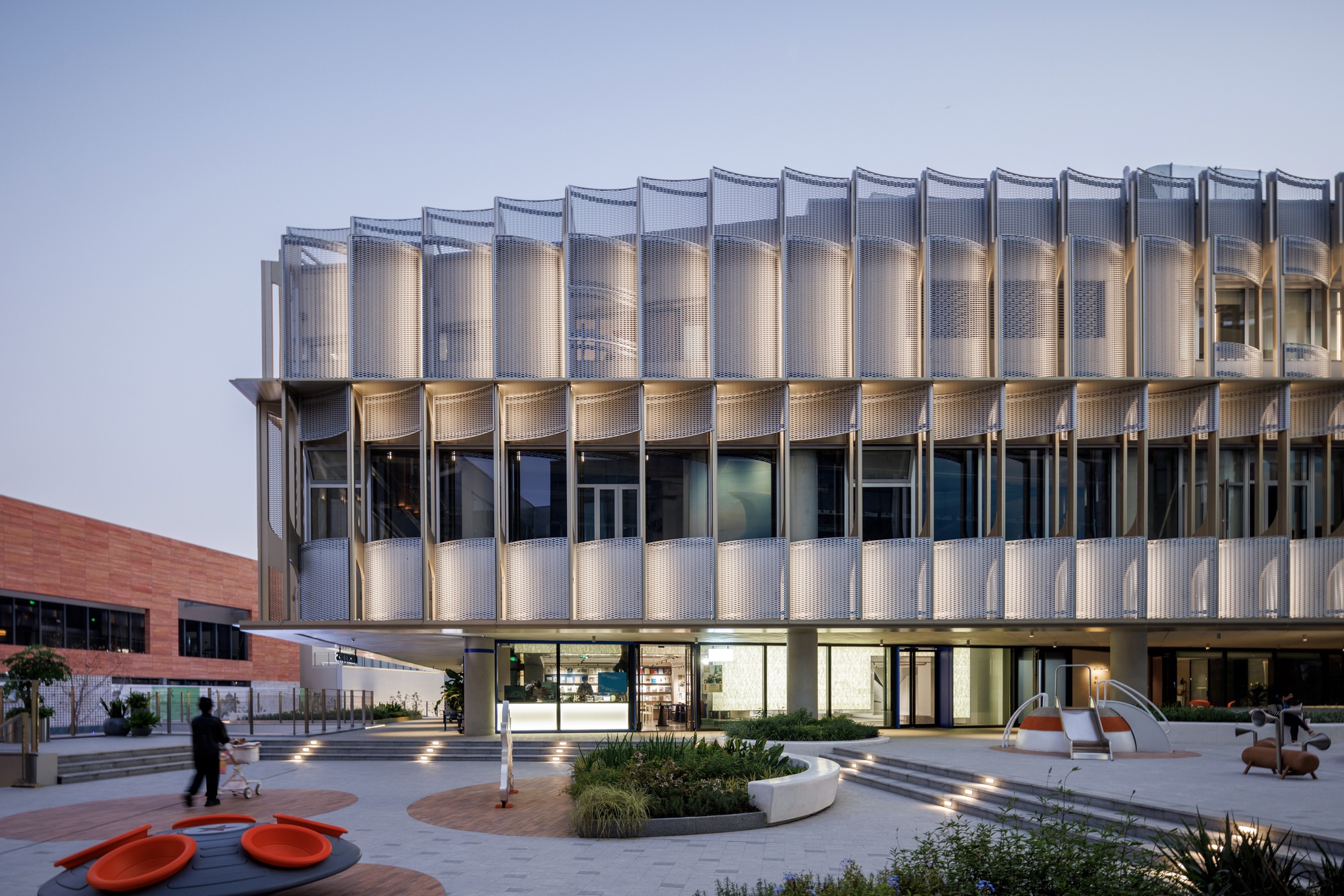 Curved white screen panels above a glazed ground floor, with orange seating and a planted bed in the plaza at twilight
