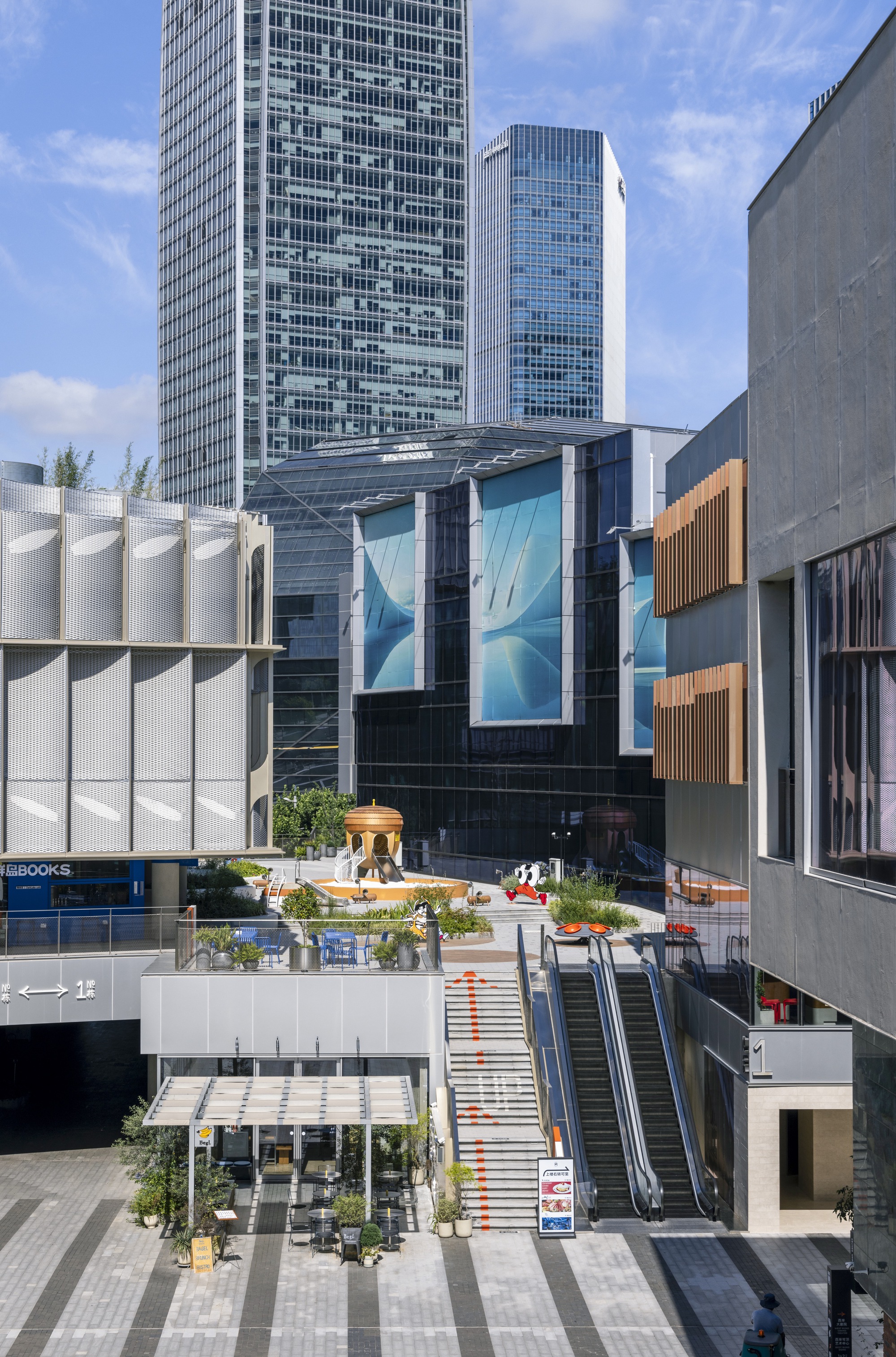 View between buildings toward a stepped plaza with escalators, a small gazebo and glass towers under a blue summer sky
