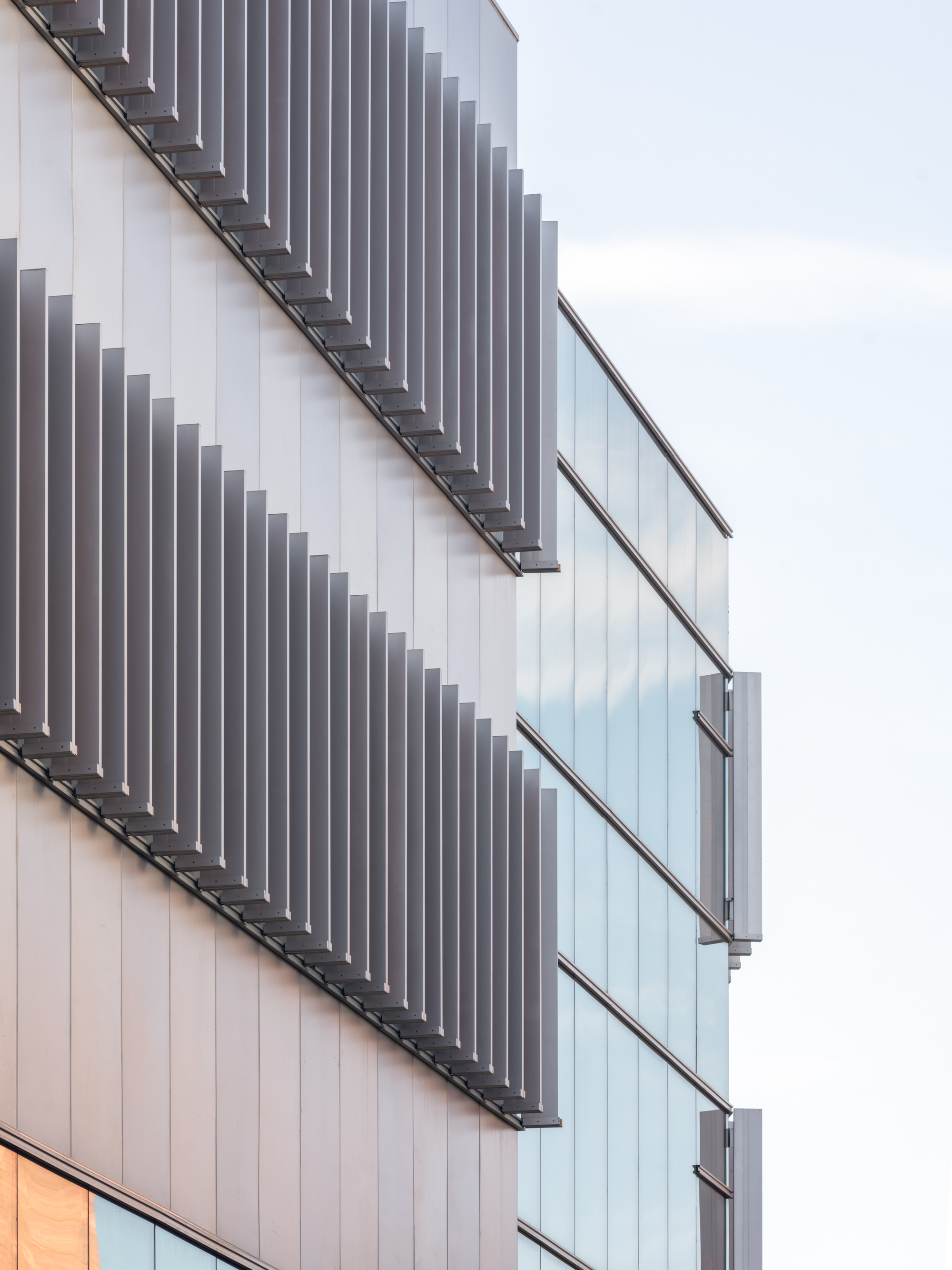 Close-up of staggered vertical metal fins on a panelled facade beside a glass curtain wall against a pale sky