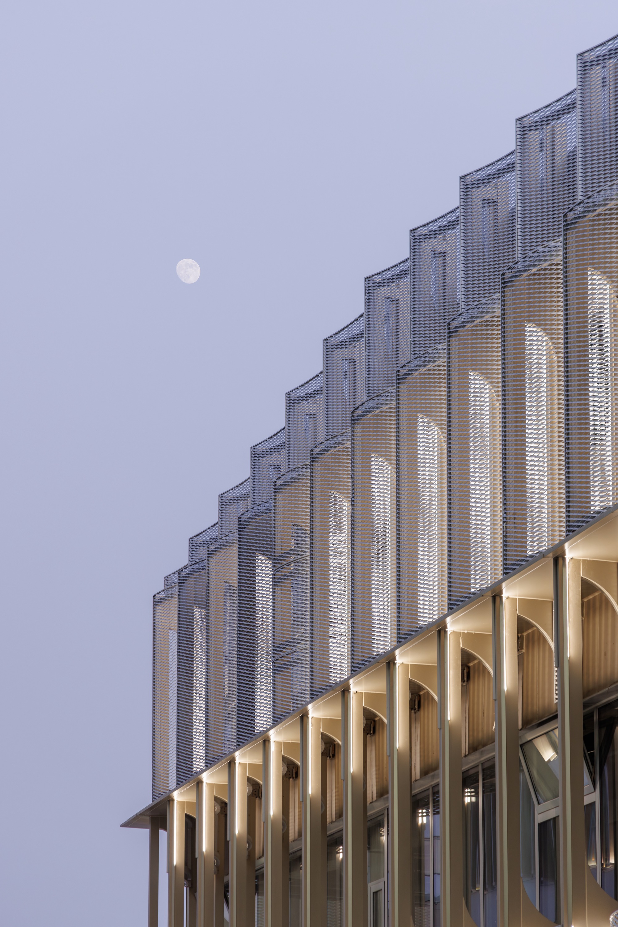 Corner of the pleated mesh screen above slender lit columns, with a pale moon in the dusk sky