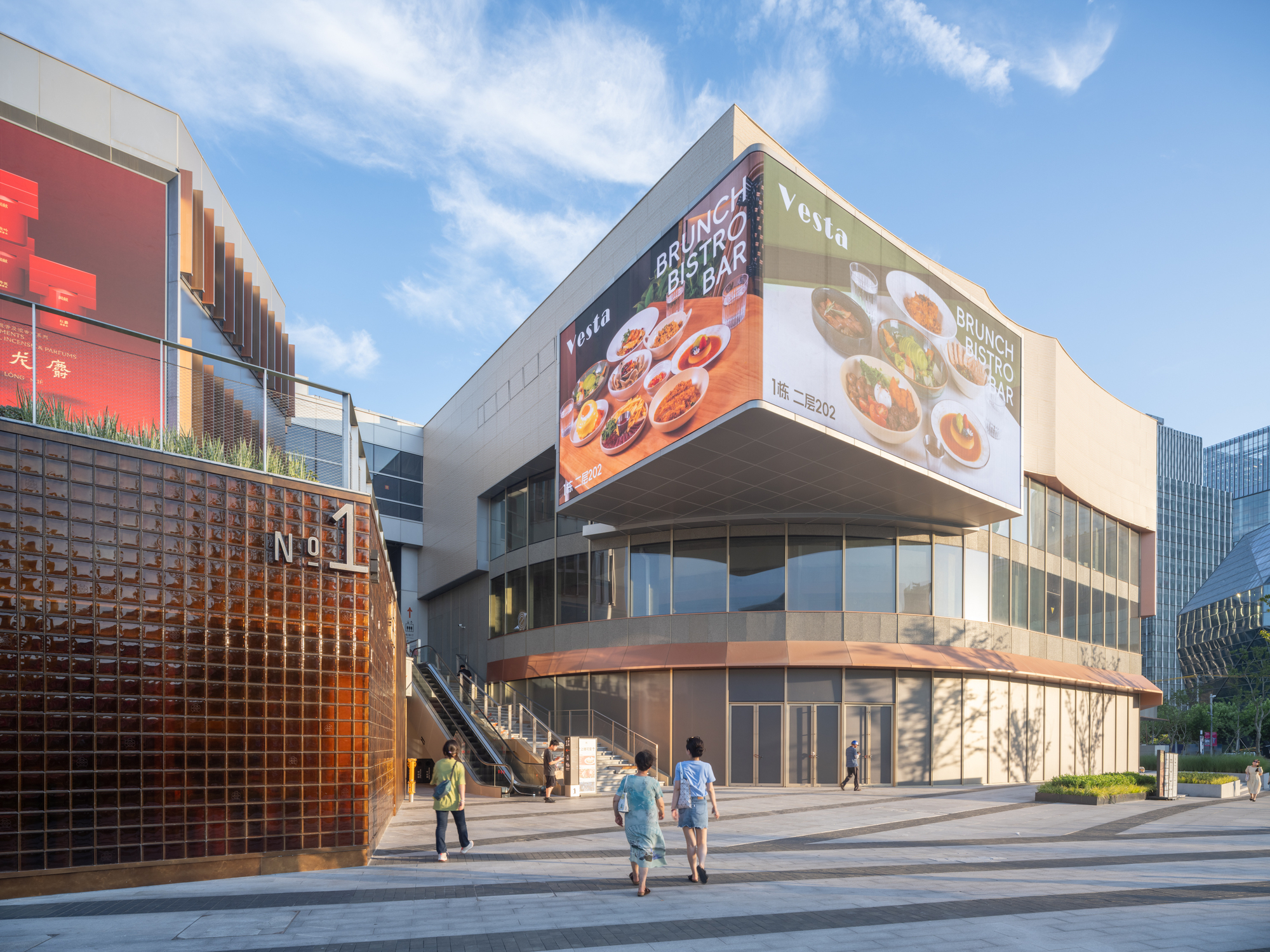 Rounded corner of a retail block with large billboard screens above, beside an amber glass-brick wall, with shoppers walking past in afternoon sun