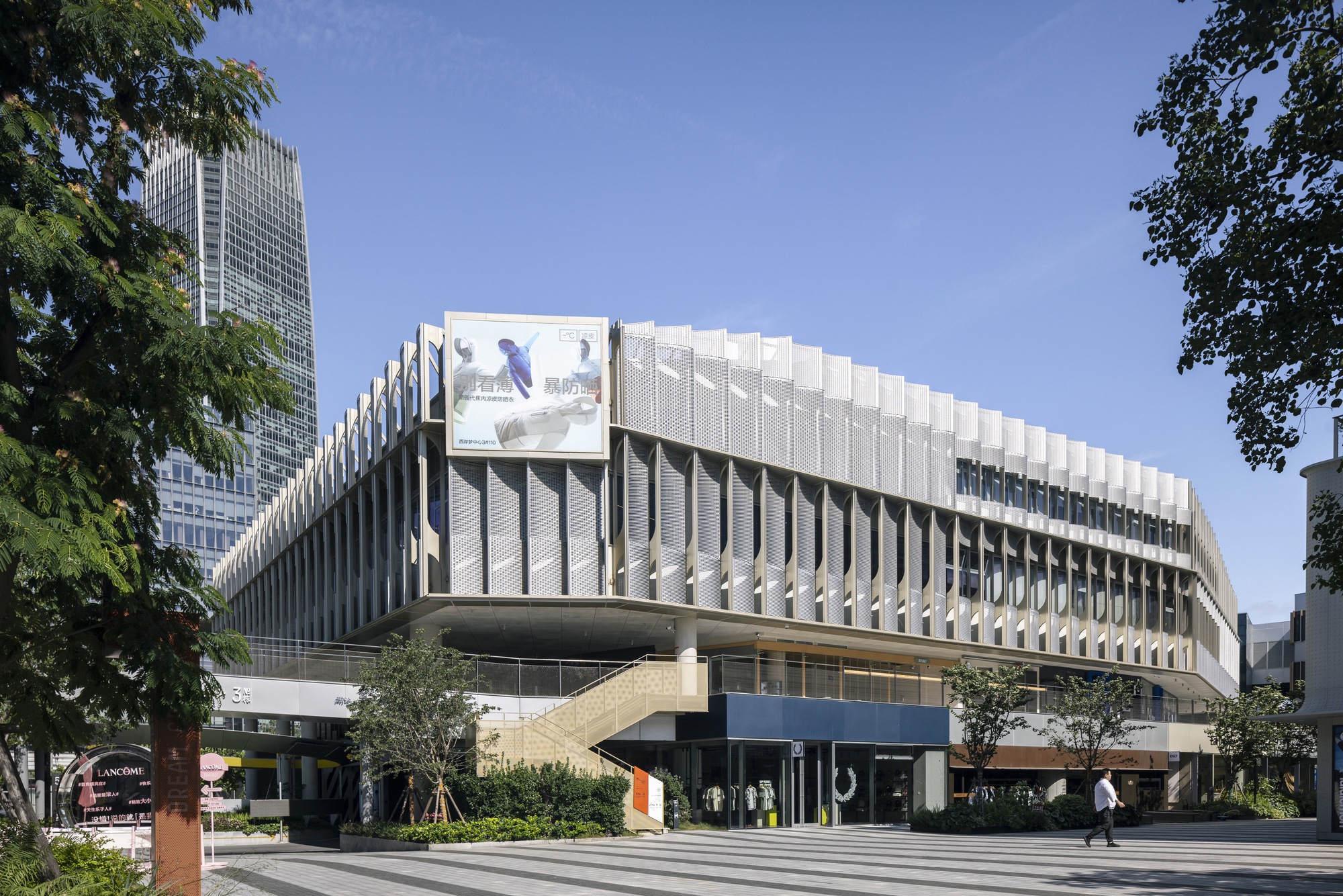Corner of a multi-storey retail building with a pleated mesh screen facade, a yellow stair and a pedestrian crossing the plaza in sunlight