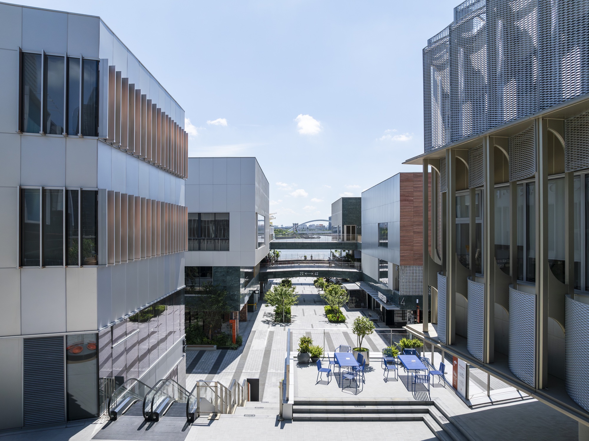 View down a pedestrian lane between white panel blocks and a perforated metal facade, with blue chairs on a terrace under clear sky