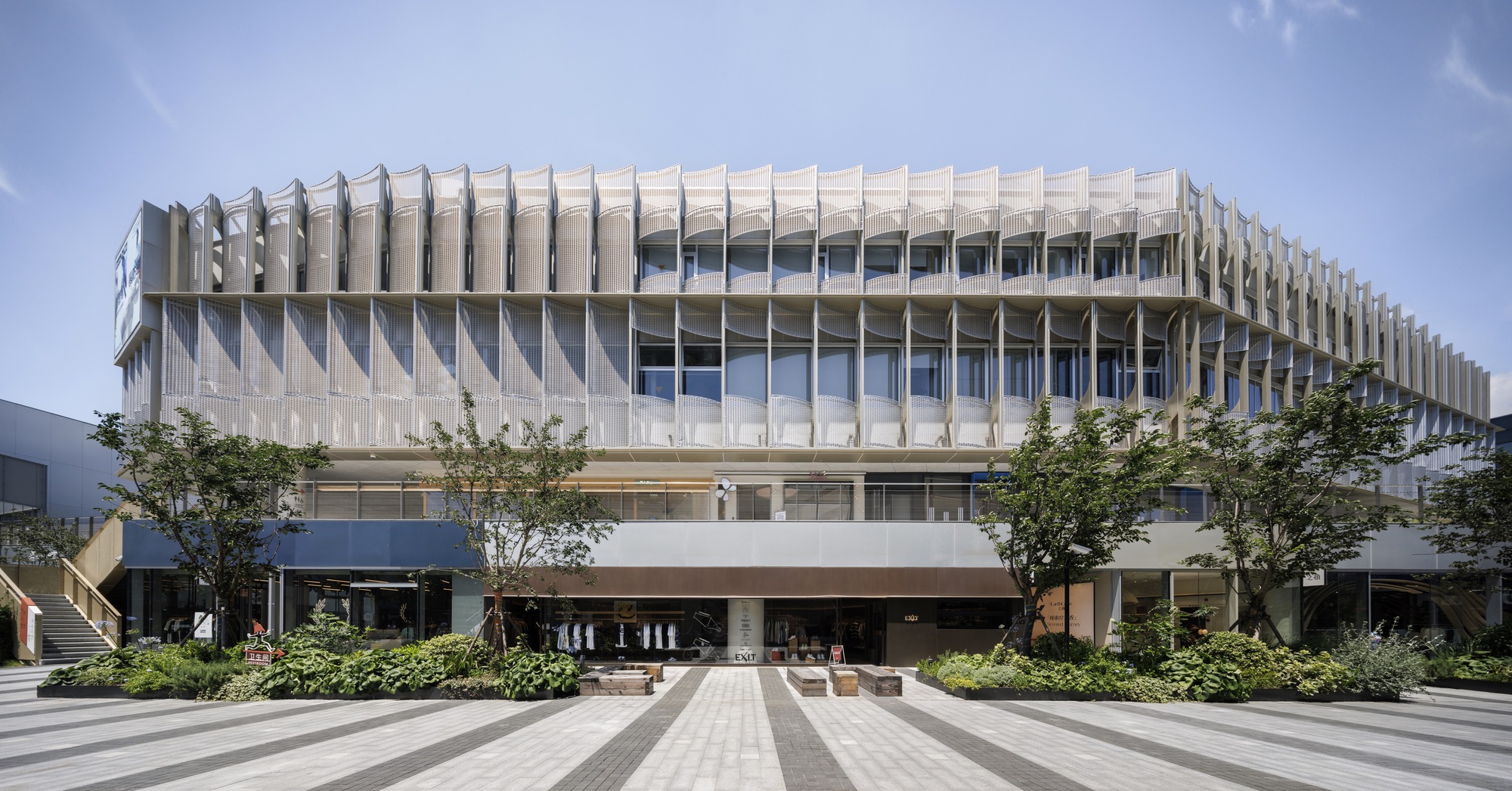 Frontal view of the curved retail facade with vertical metal fins above a paved plaza and planted beds with young trees