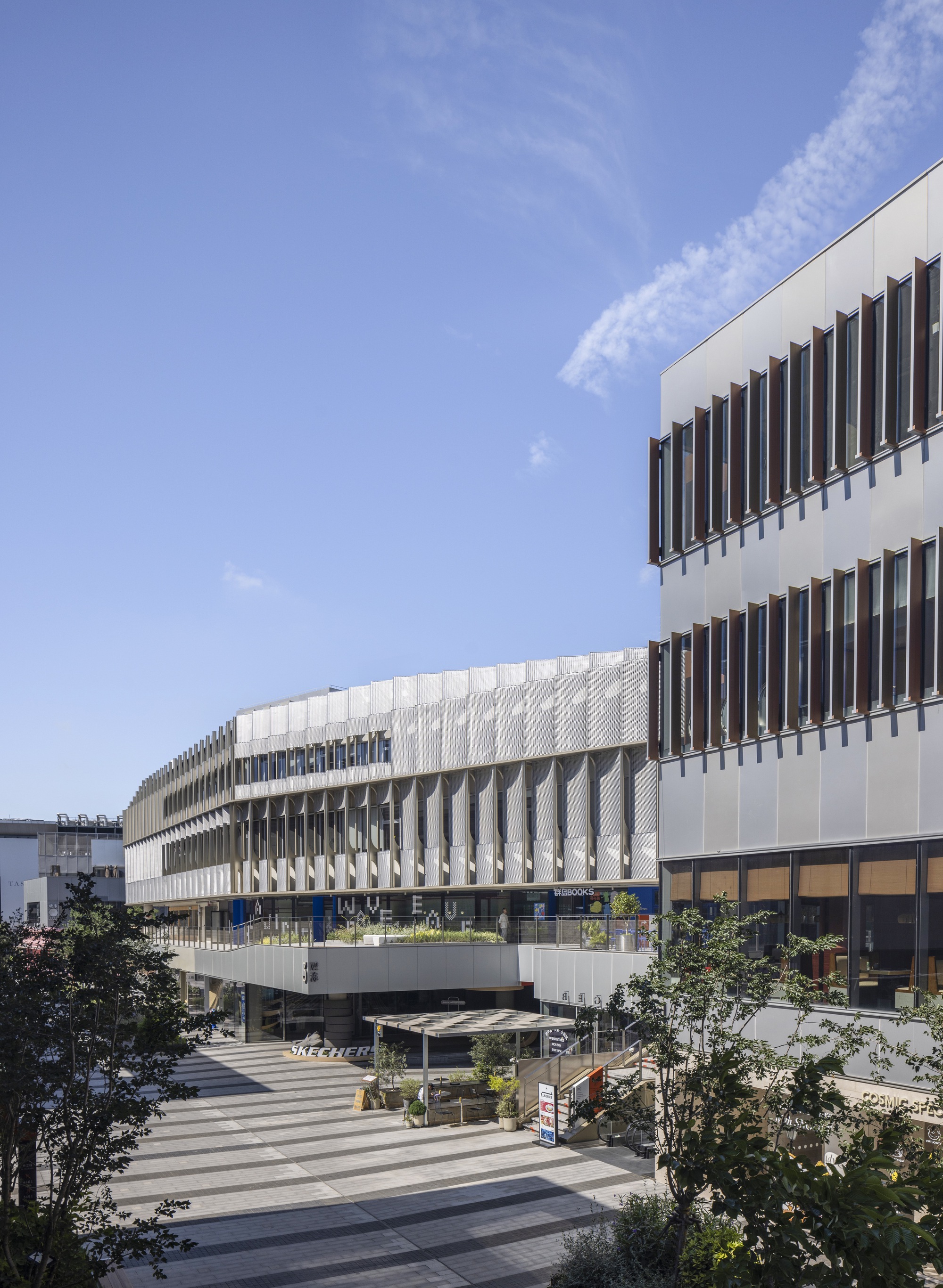 Curving facade of fin-screened balconies seen across a striped paved plaza, beside a block with rust-colored vertical louvers under a blue sky