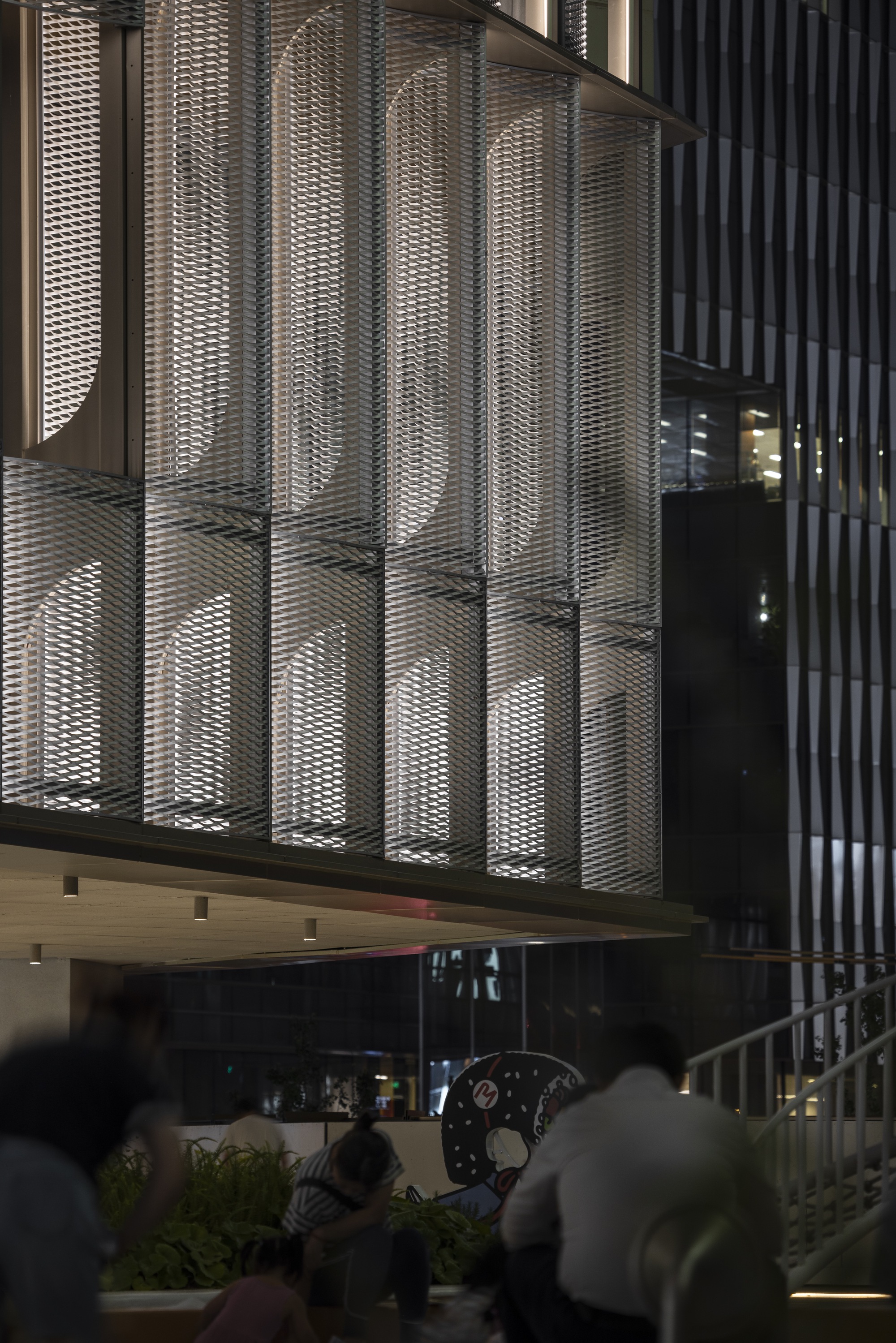 Night view of expanded metal mesh screens with arched openings above a planted seating area where people sit