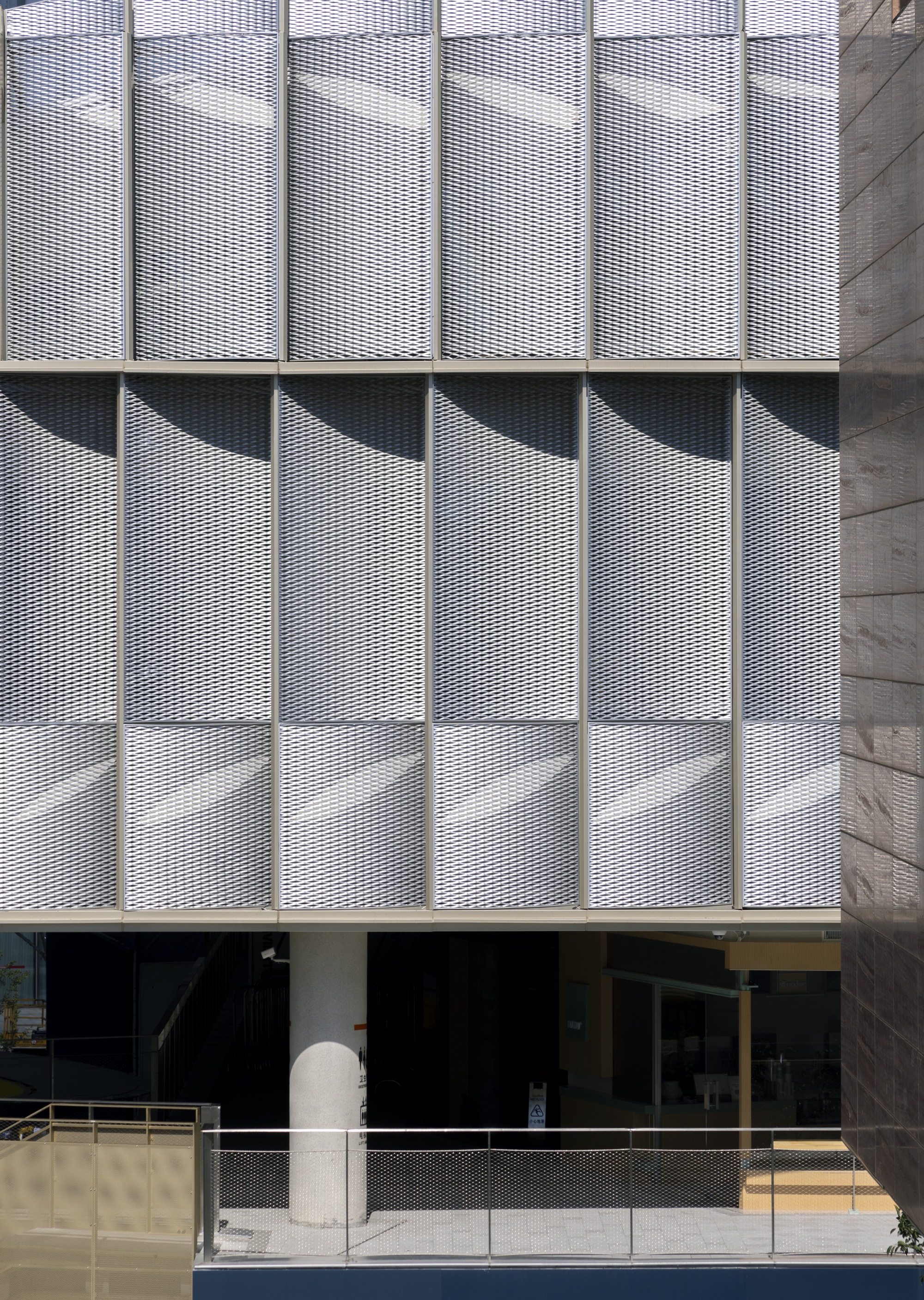 Facade of expanded metal mesh panels casting angled shadows above a round concrete column in bright midday sun