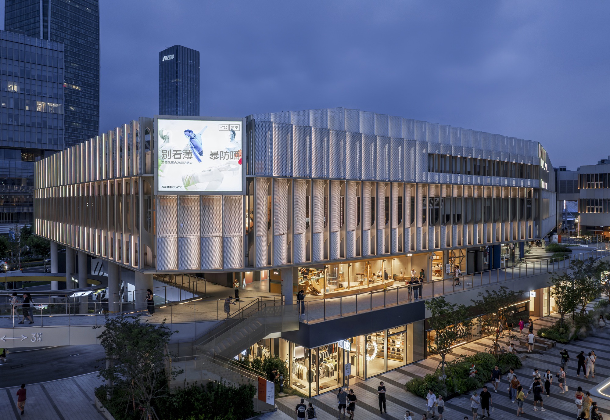 Elevated dusk view of the pleated mesh-clad retail building with an illuminated screen and shoppers on the stepped plaza