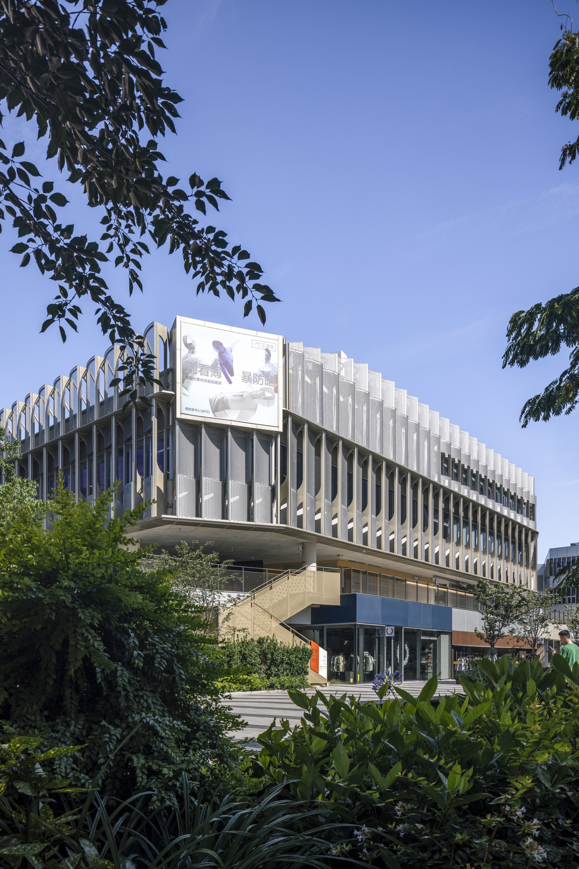 Corner of the mesh-screened building with a billboard, seen through leafy branches under a clear blue sky