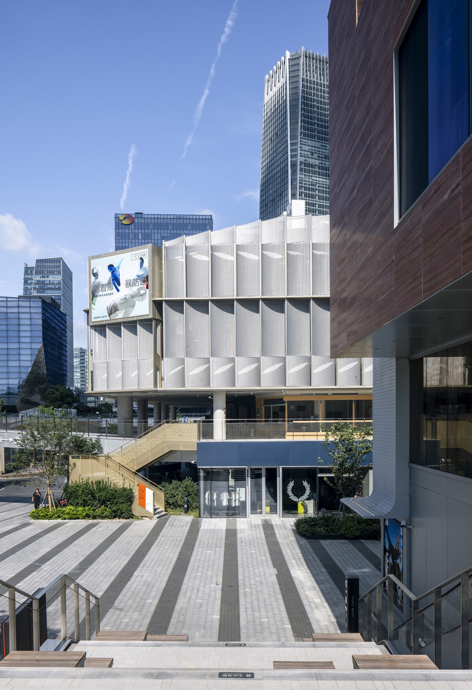 Paved plaza with dark stripes leading to the screened building and a staircase, framed by towers on a sunny day