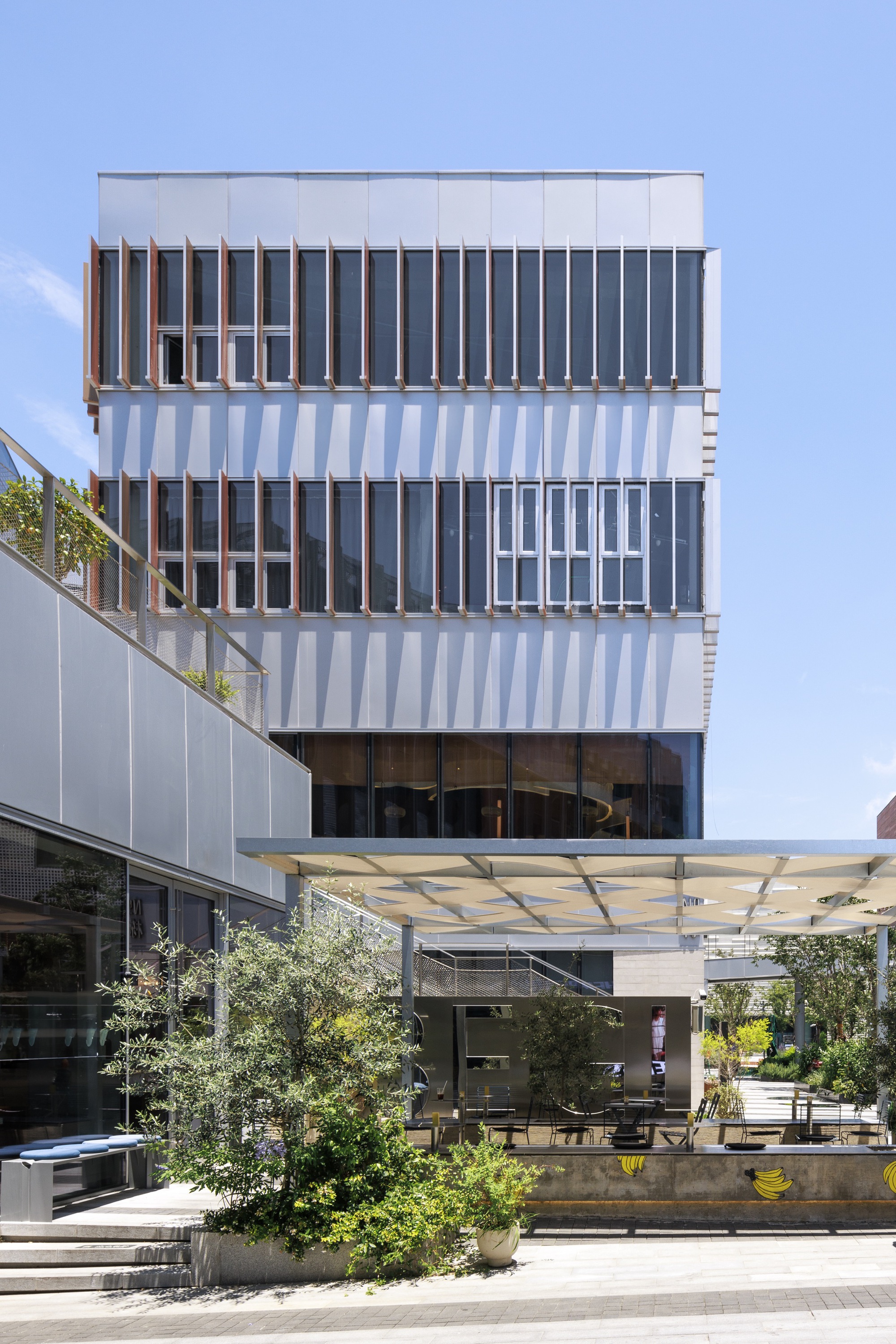 Stepped facade of white metal panels and copper-toned vertical fins behind a planted terrace and canopy under a clear blue sky