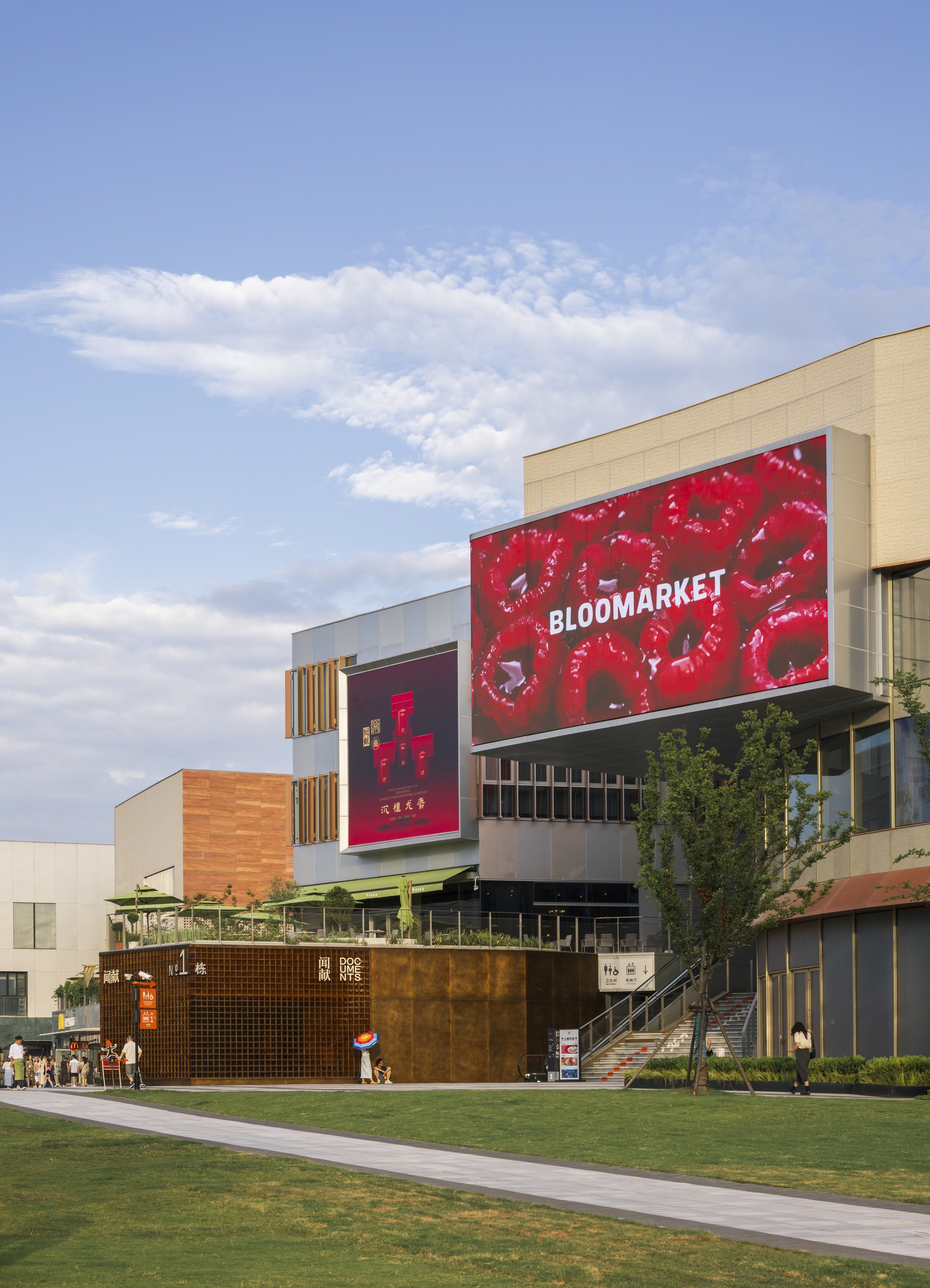 Large LED screen cantilevering from a pale stone block above a lawn, with a rusted metal pavilion and people strolling