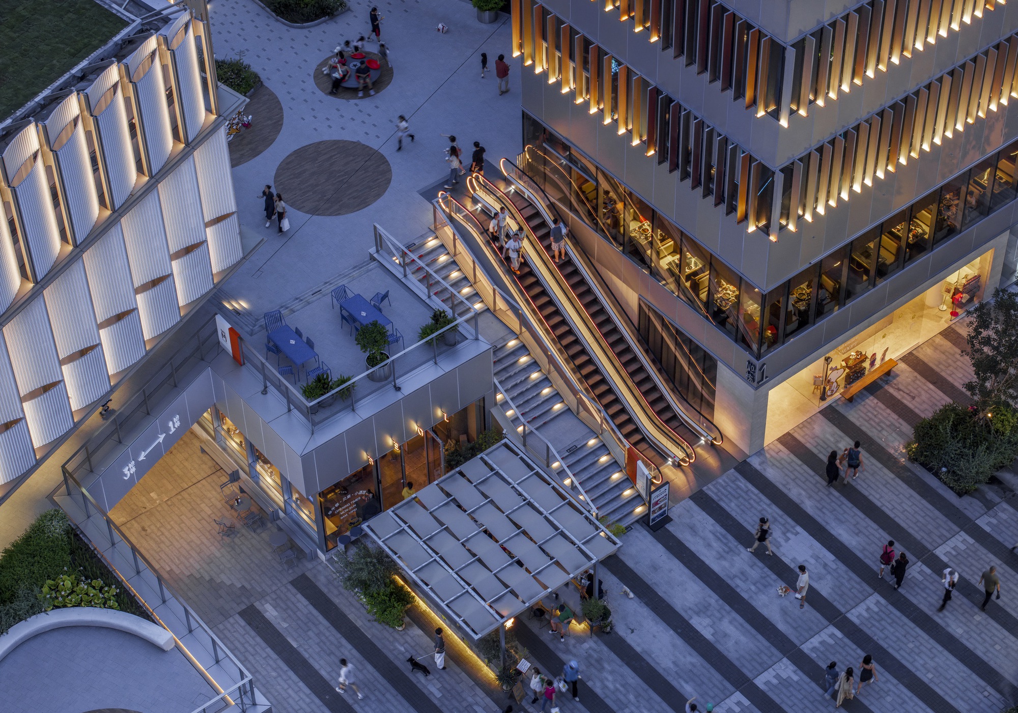 Aerial view of escalators and stairs rising from a striped granite plaza at dusk, with lit fins and people crossing