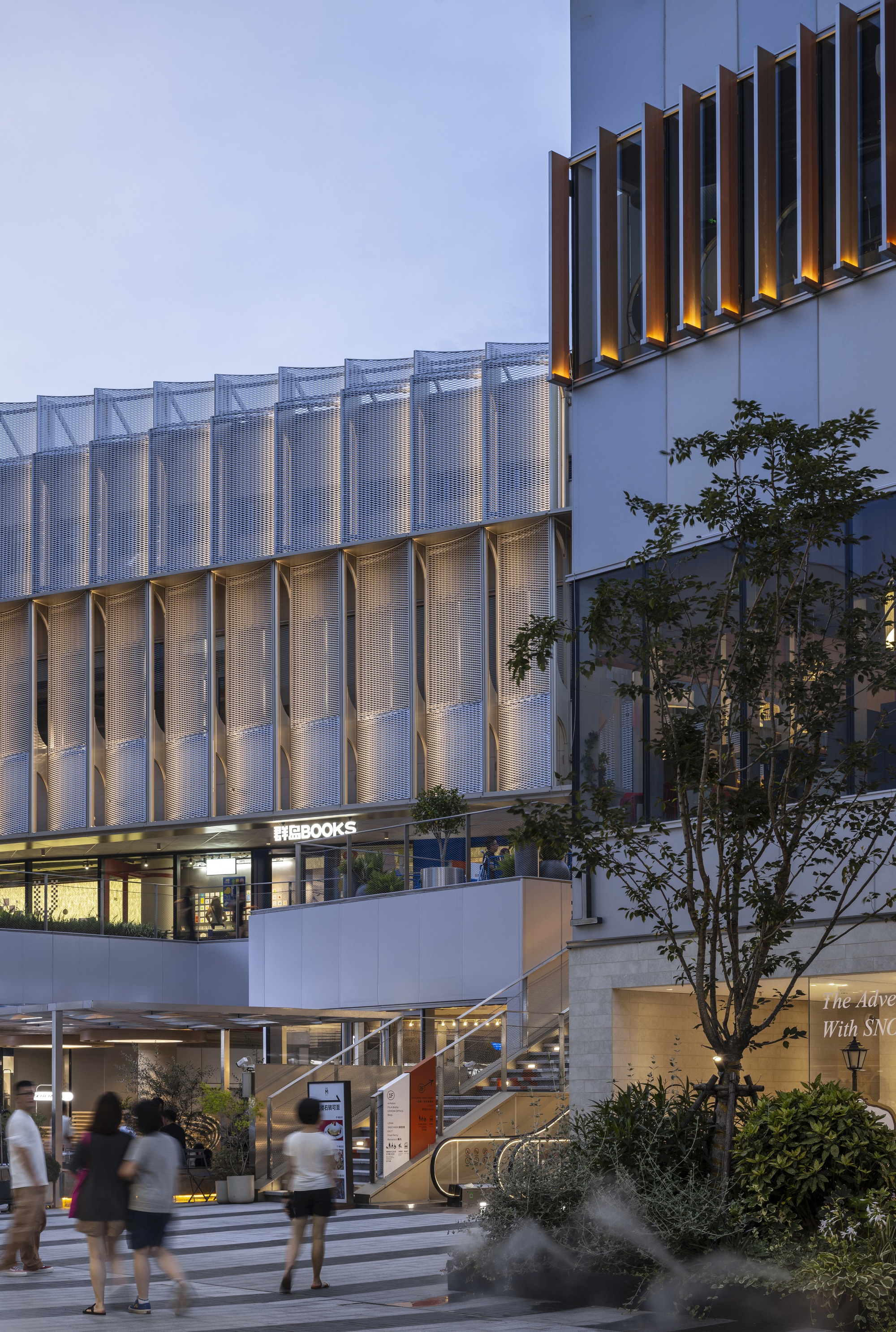 Translucent curved mesh screens wrapping a bookstore facade beside a fin-clad tower, with a tree and walkers at dusk