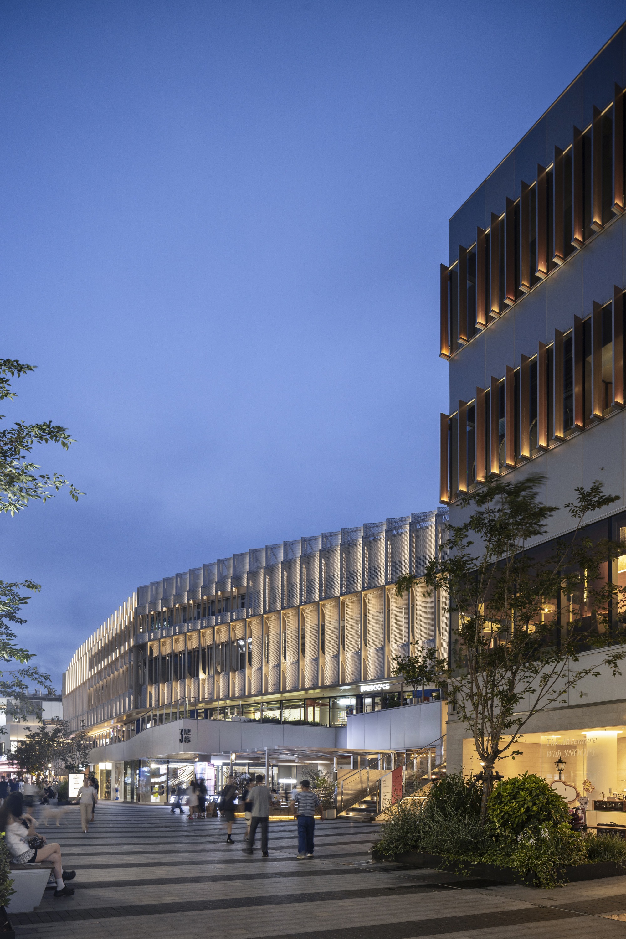 Plaza at blue hour with a long mesh-clad podium glowing beside a tower with copper fins, pedestrians and a tree in front