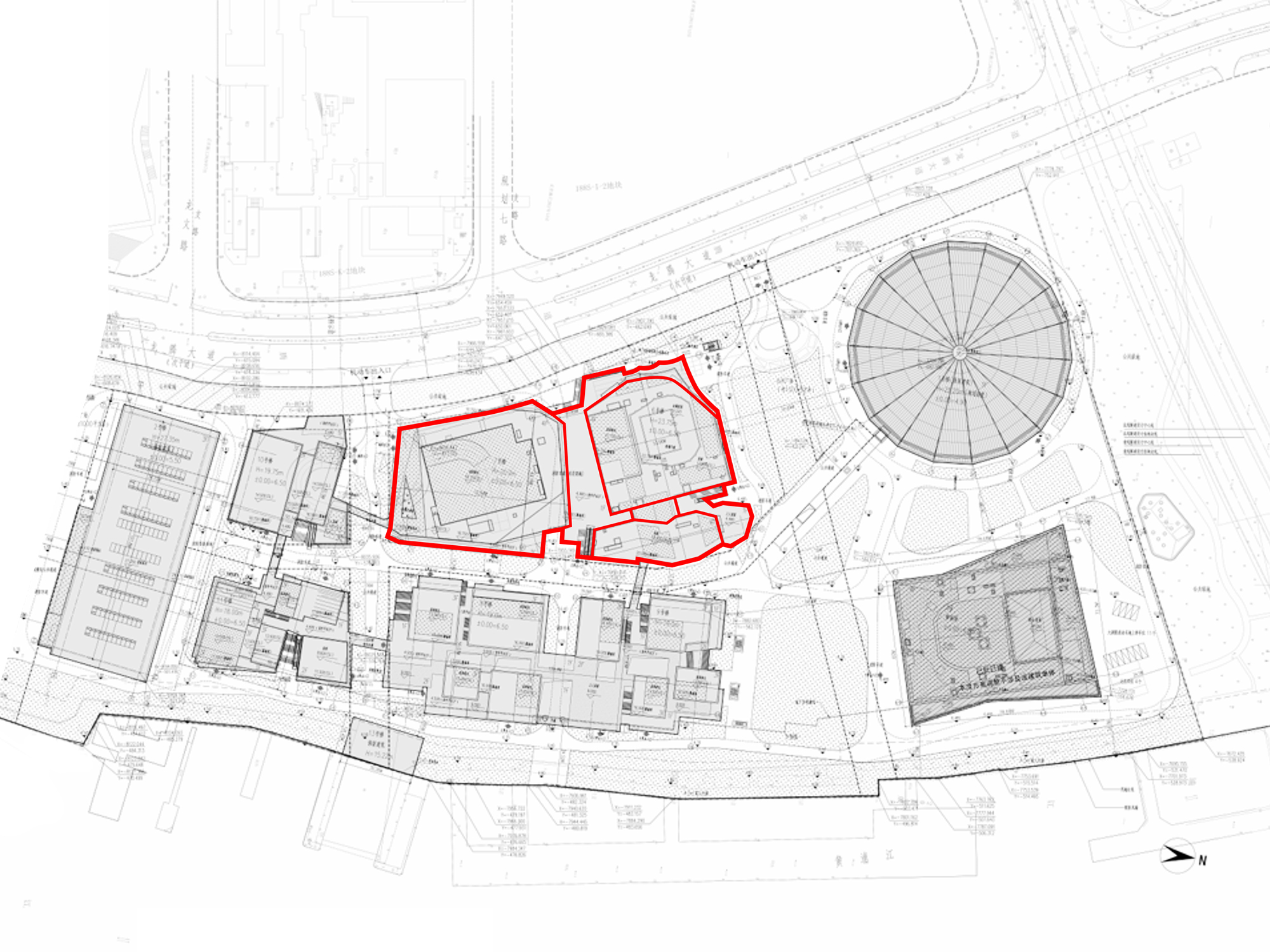 Site plan drawing with three irregular building footprints outlined in red beside a circular ribbed-roof structure and surrounding blocks along a waterfront edg