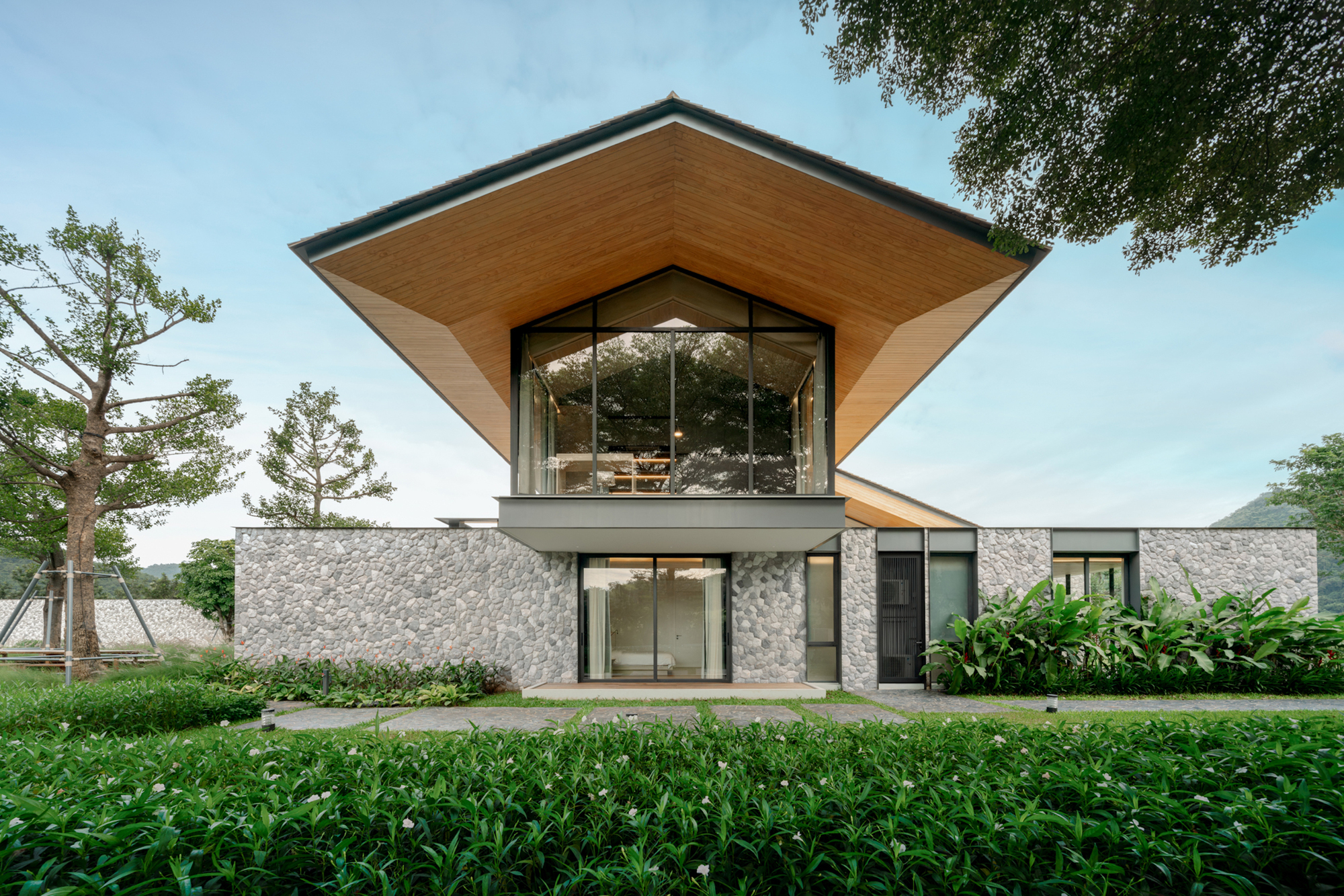 Front facade with a wide timber-lined gabled roof over a river stone wall, framed by tall trees and flowering shrubs