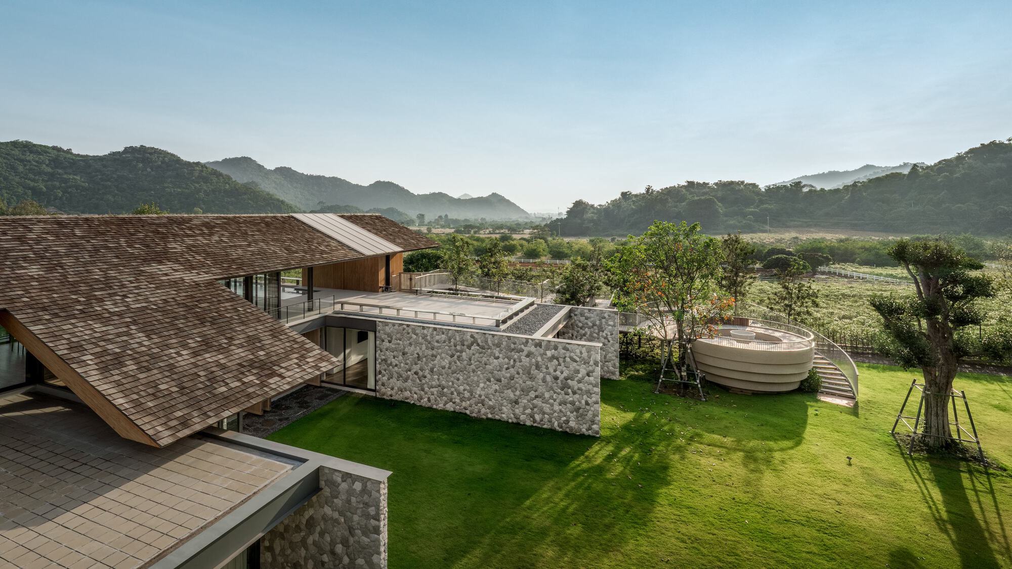 Elevated view of shingled roofs and a stone-clad wall stepping down to a lawn, with forested hills in morning haze