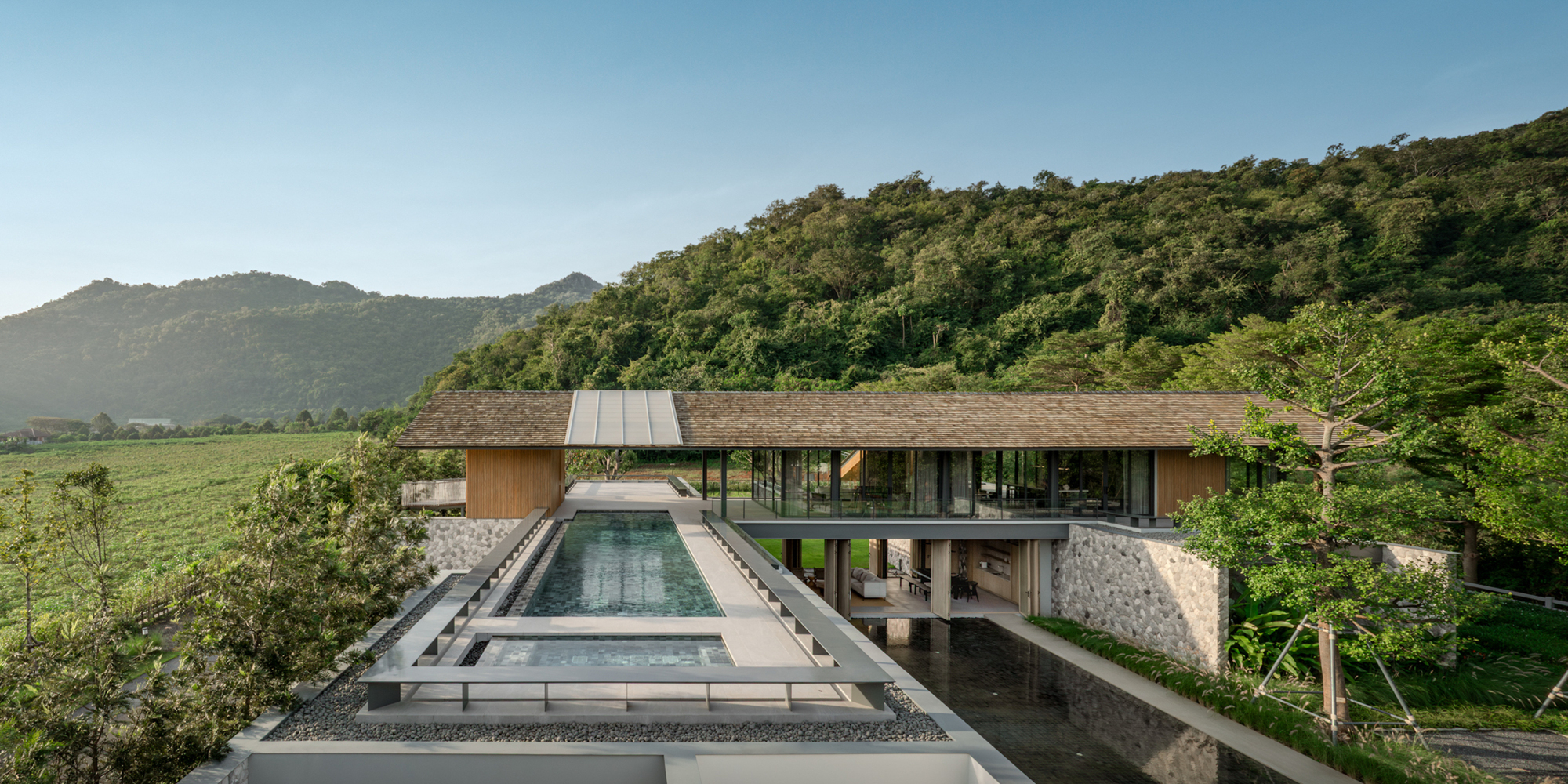 Long pavilion with a low shingle roof above a rooftop pool and reflecting water, set against a forested hillside