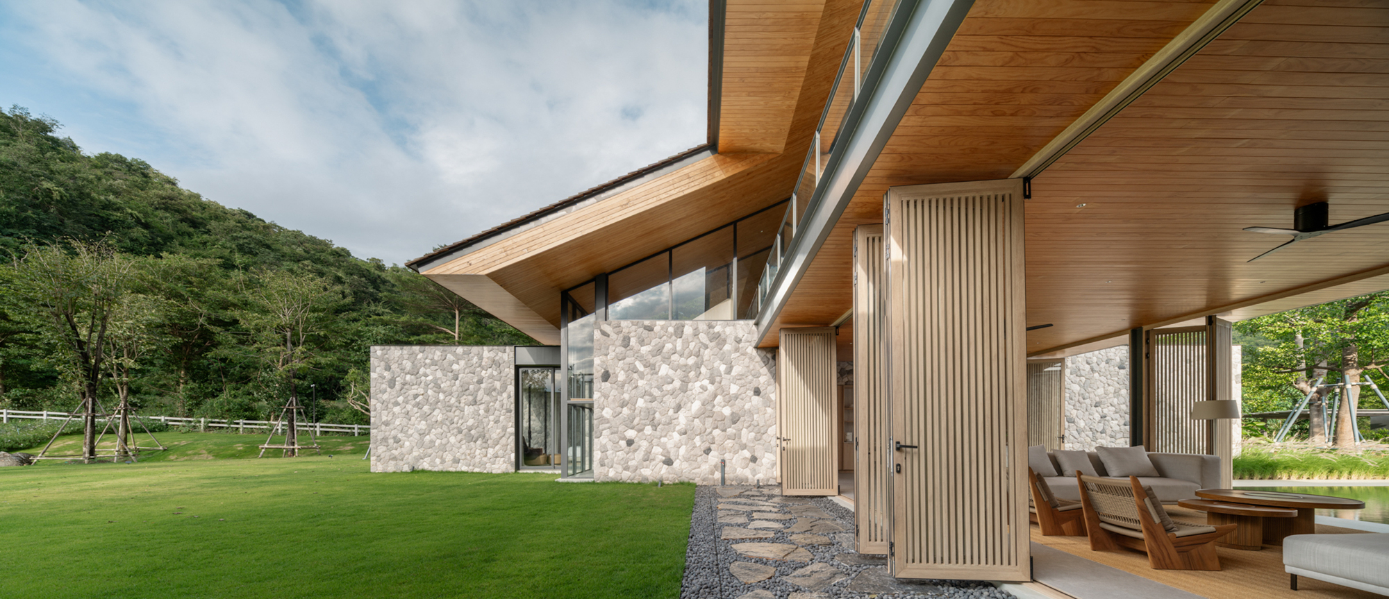 Folding timber slatted doors open onto a covered terrace with lounge seating, beside stone walls and a sunlit lawn