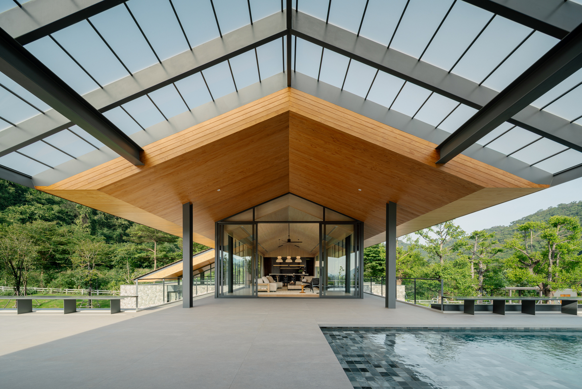 Translucent canopy roof on steel beams above a paved terrace and pool, with a timber soffit leading to the glazed living room