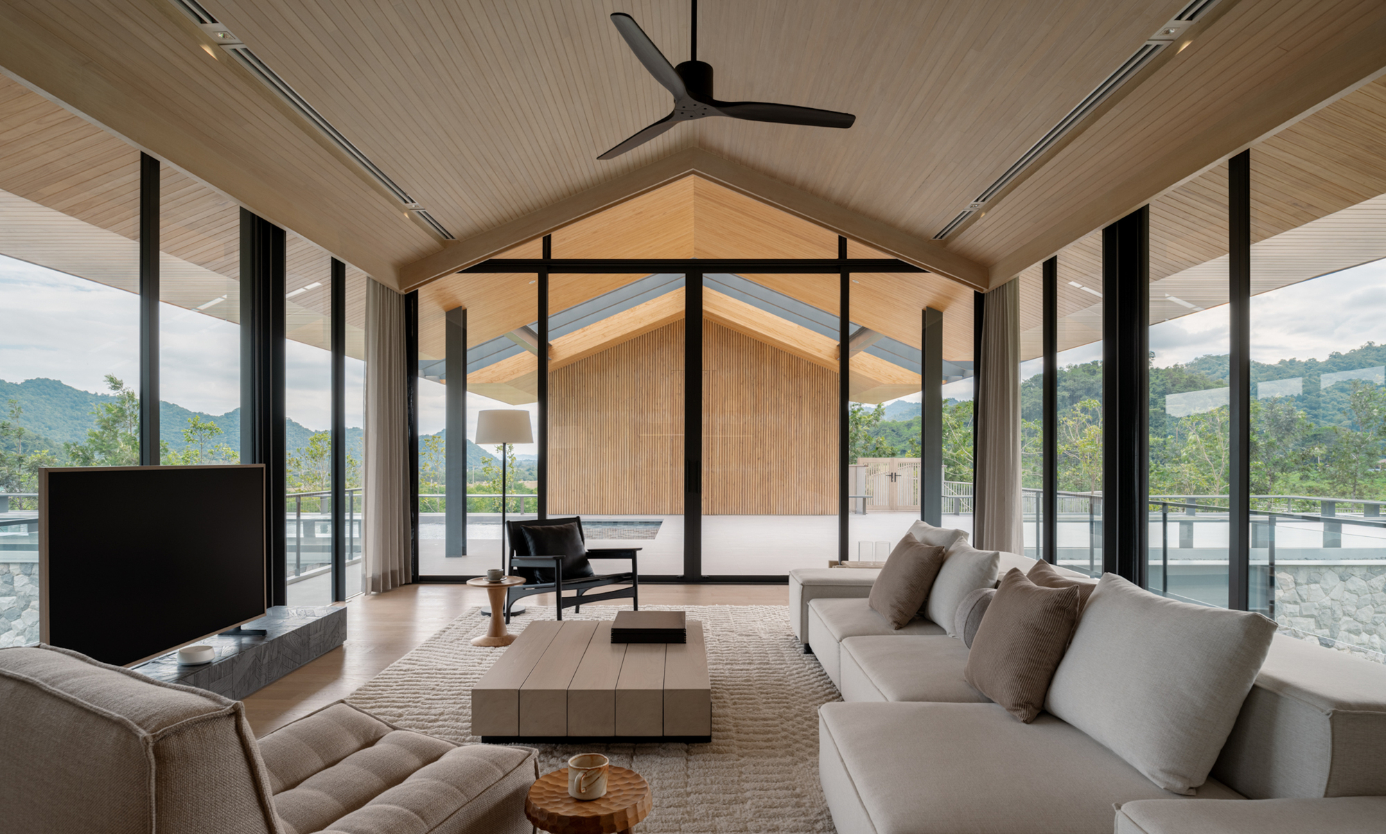 Upper living room with a pitched timber ceiling, ceiling fan, and sofa group facing floor-to-ceiling glass and cloudy green hills
